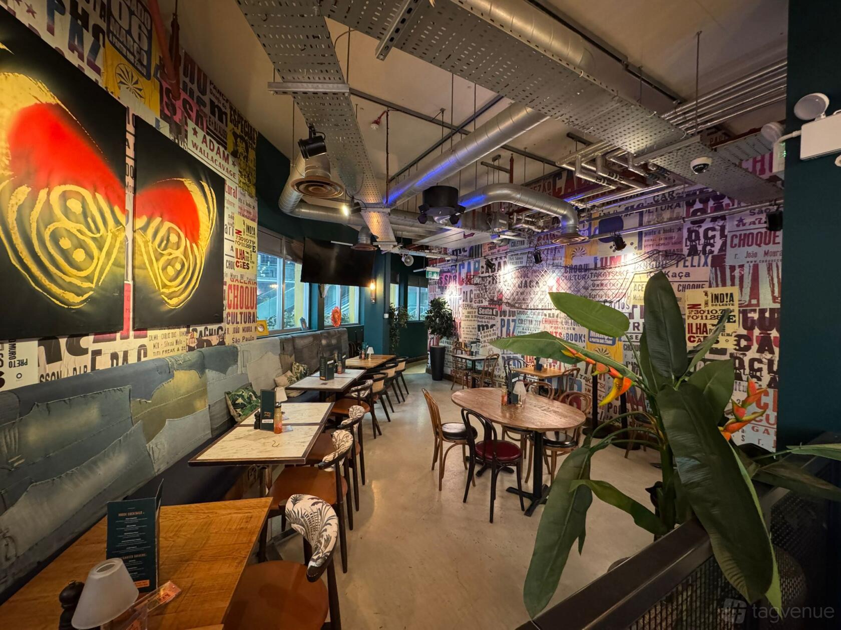 An event space in a restaurant with colorful mural walls, exposed ductwork, and mixed wooden tables at Cabana at The O2.