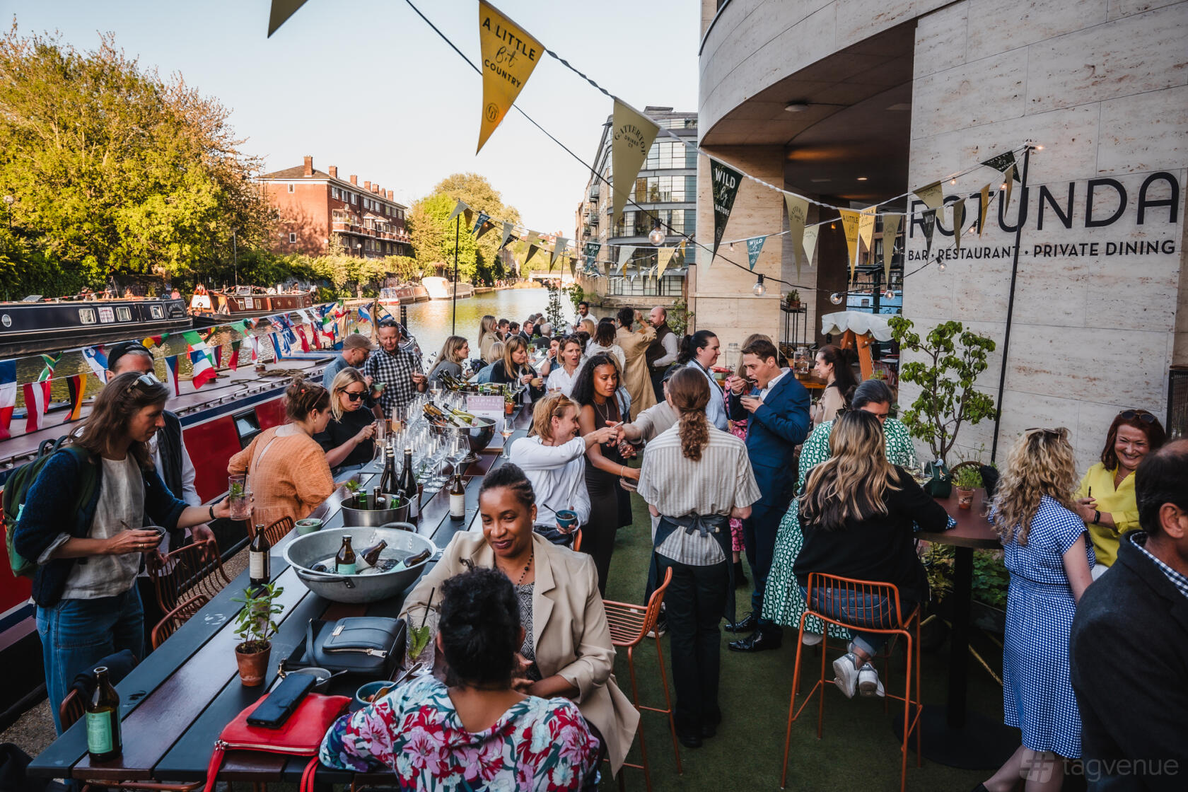 An outdoor restaurant area with communal tables beside a canal and festive bunting at Rotunda Bar and Restaurant.
