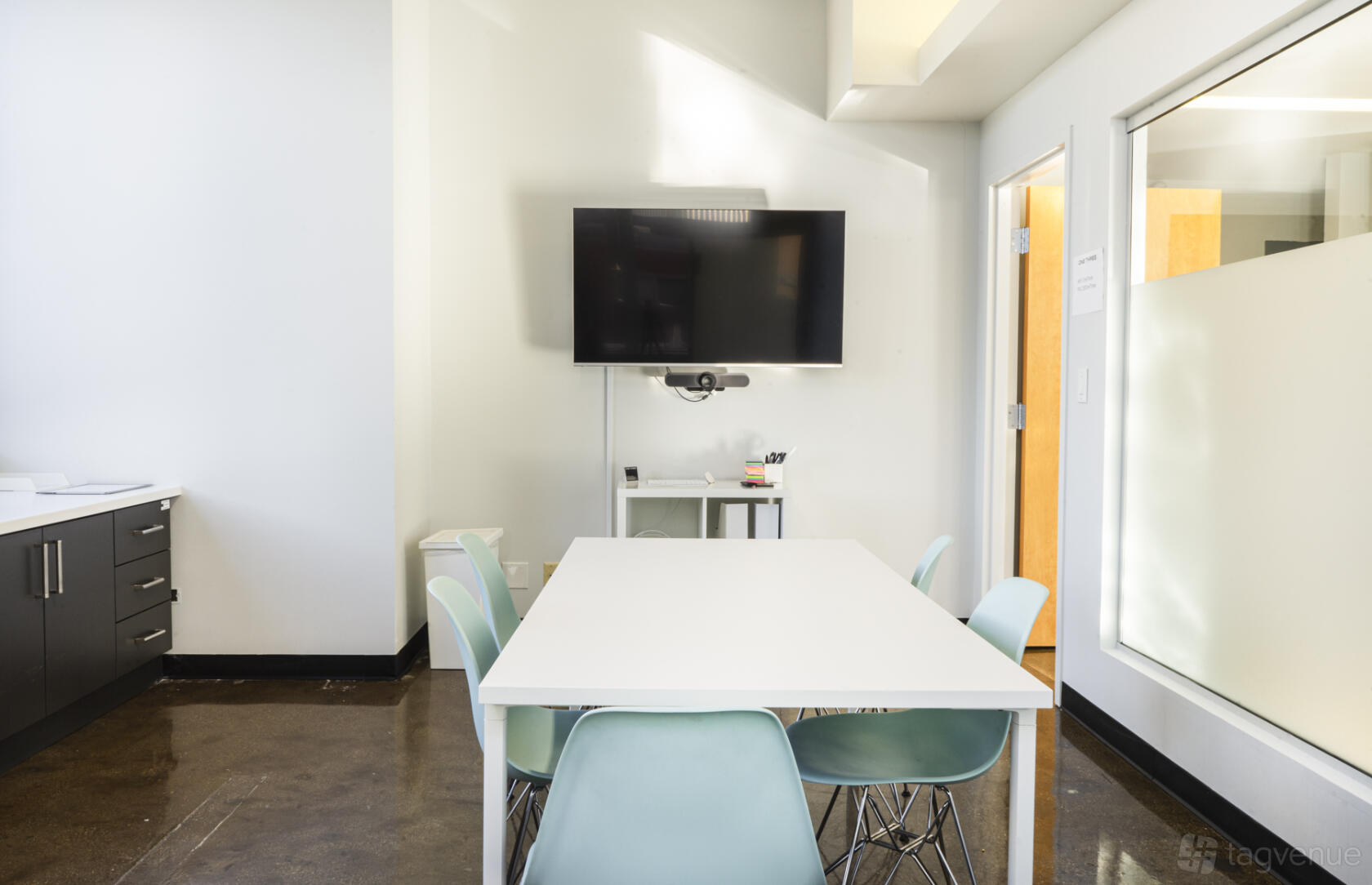 A meeting room with a white table, teal chairs, wall-mounted TV, and glass partition at One Three Workspace.