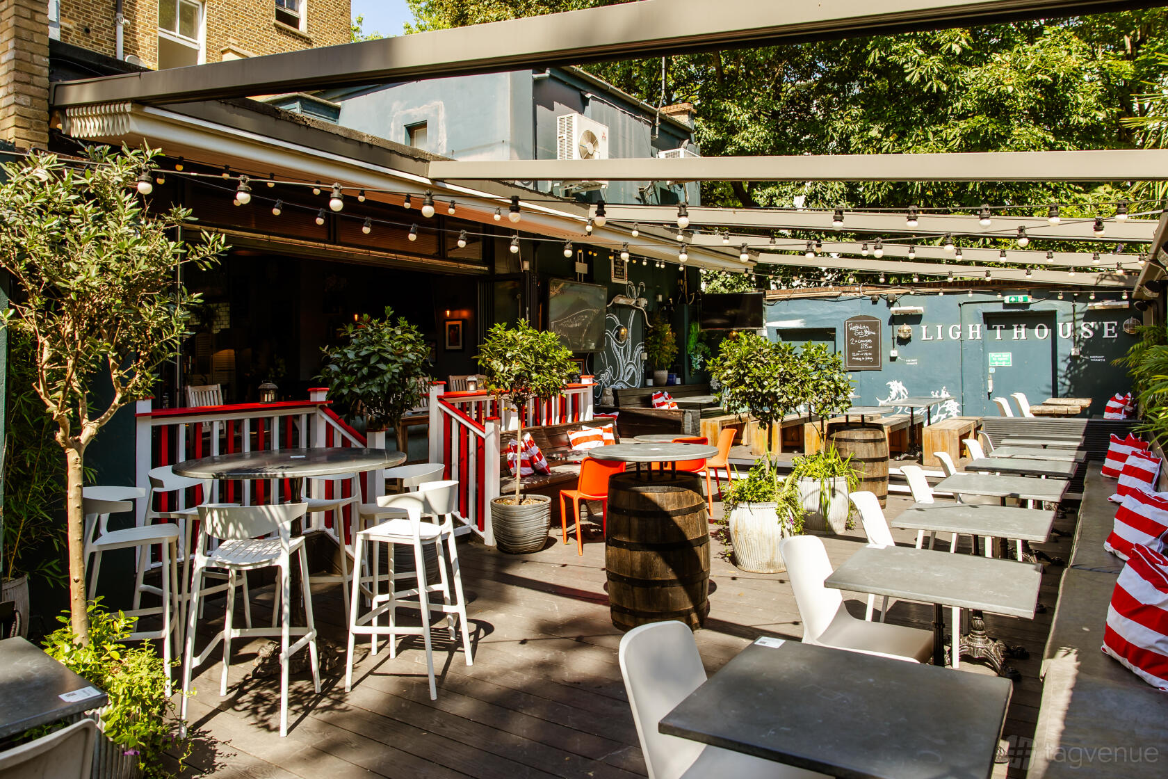 An outdoor pub space with wooden decking, potted plants, striped seating, and string lights at The Lighthouse.