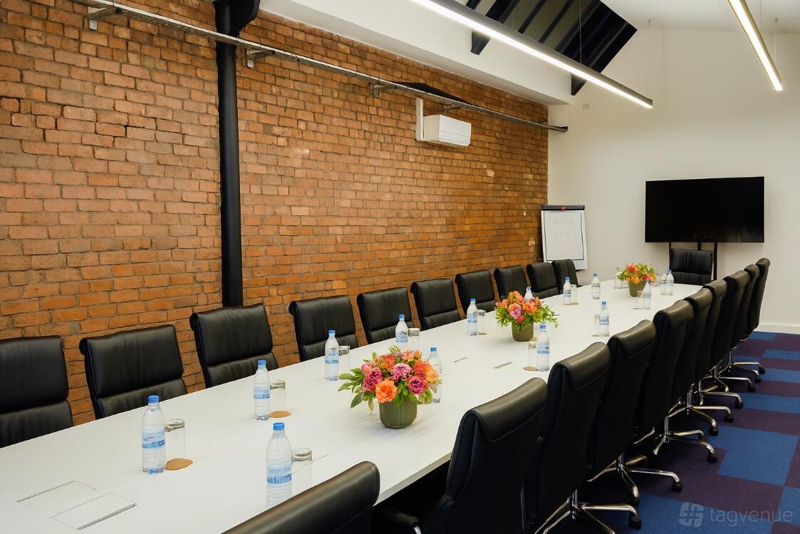 A meeting room with exposed brick walls, a long white conference table, and black chairs at Heritage 1907- Ram Mill.