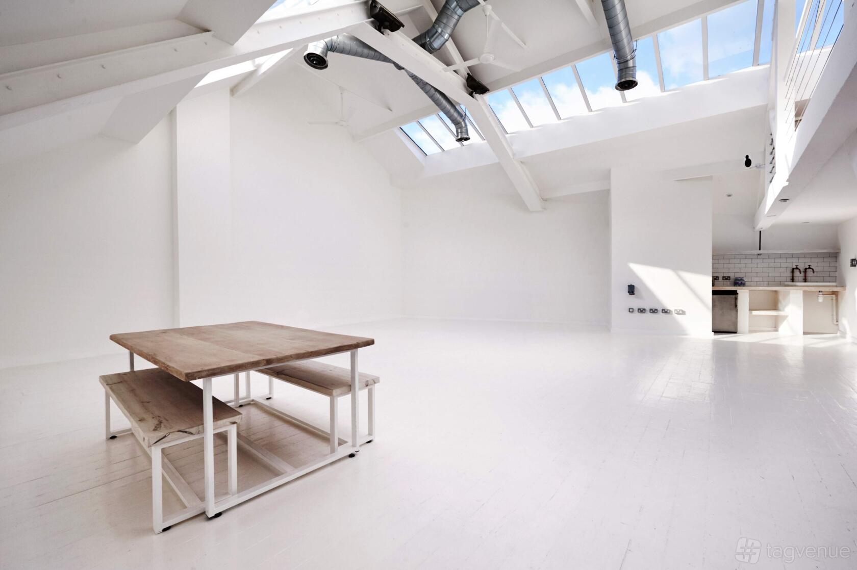 An event space with skylights, exposed beams, white floors, and a wooden table with benches at fivefourstudios.
