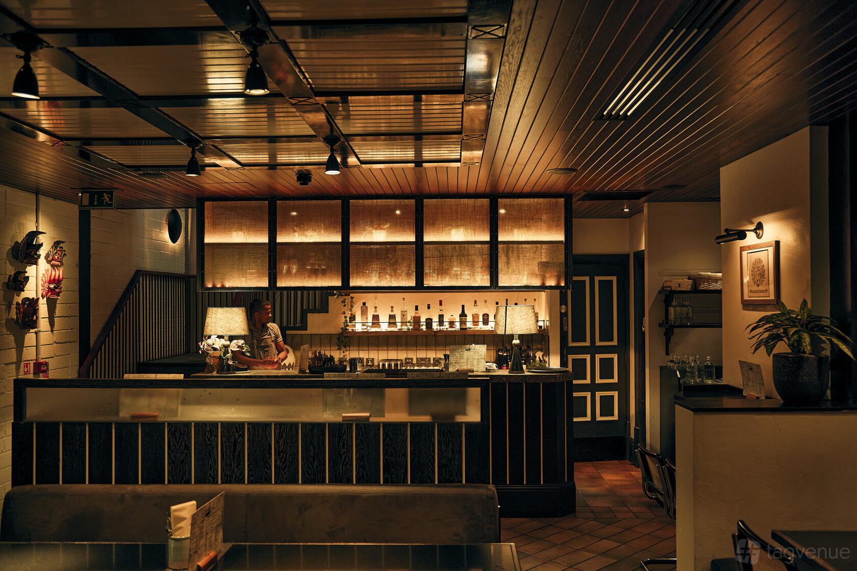 A restaurant interior with warm lighting, a wooden ceiling, backlit shelves of bottles, and a bar counter at Hoppers Marylebone.