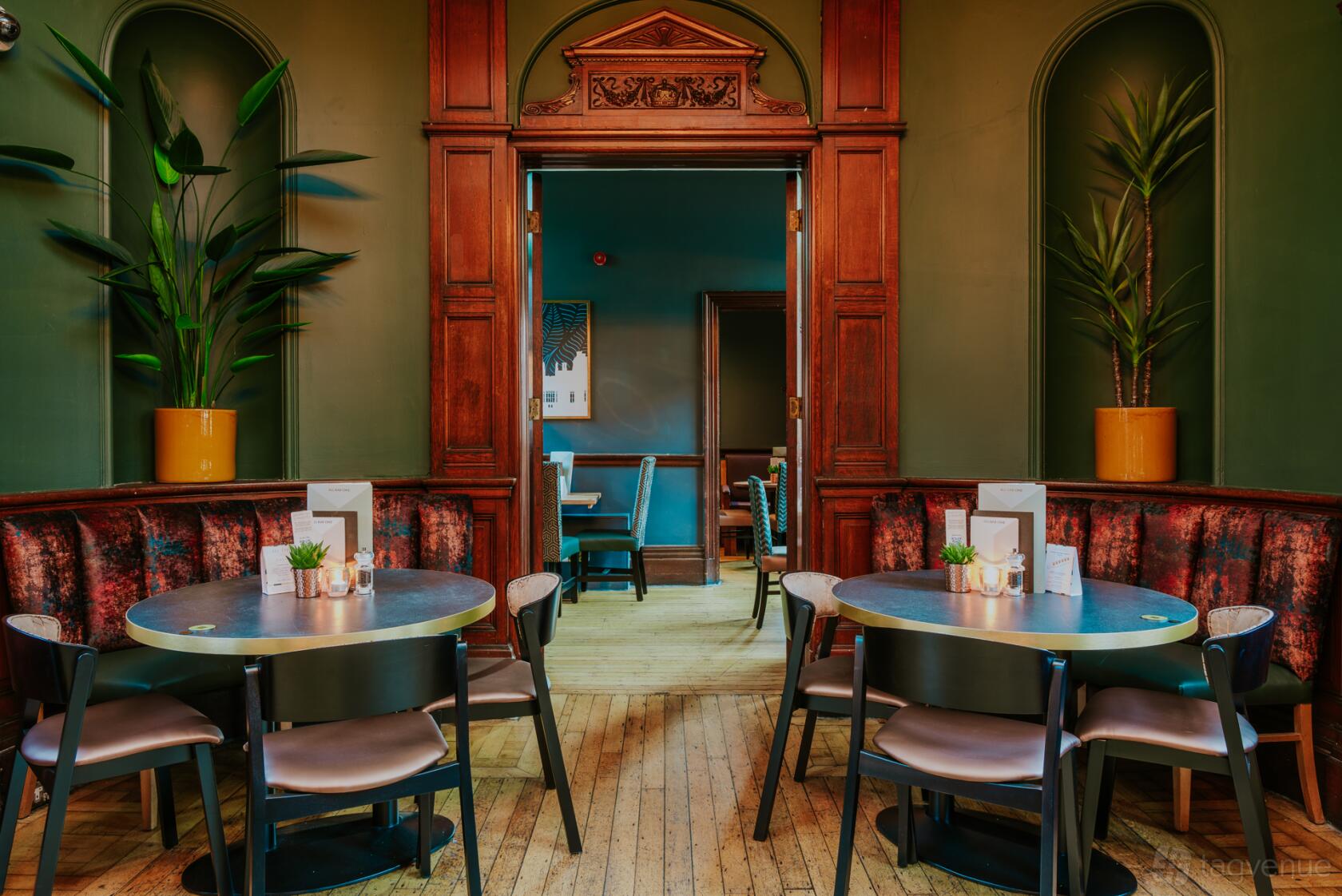 A bar with round tables, plush banquette seating, wood panel accents, and potted plants at All Bar One Windsor.