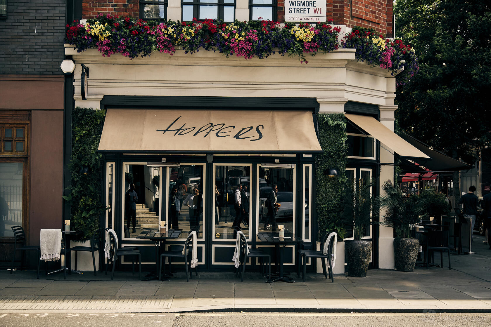 A street-facing restaurant with large windows, tan awnings, and hanging flowers at Hoppers Marylebone.