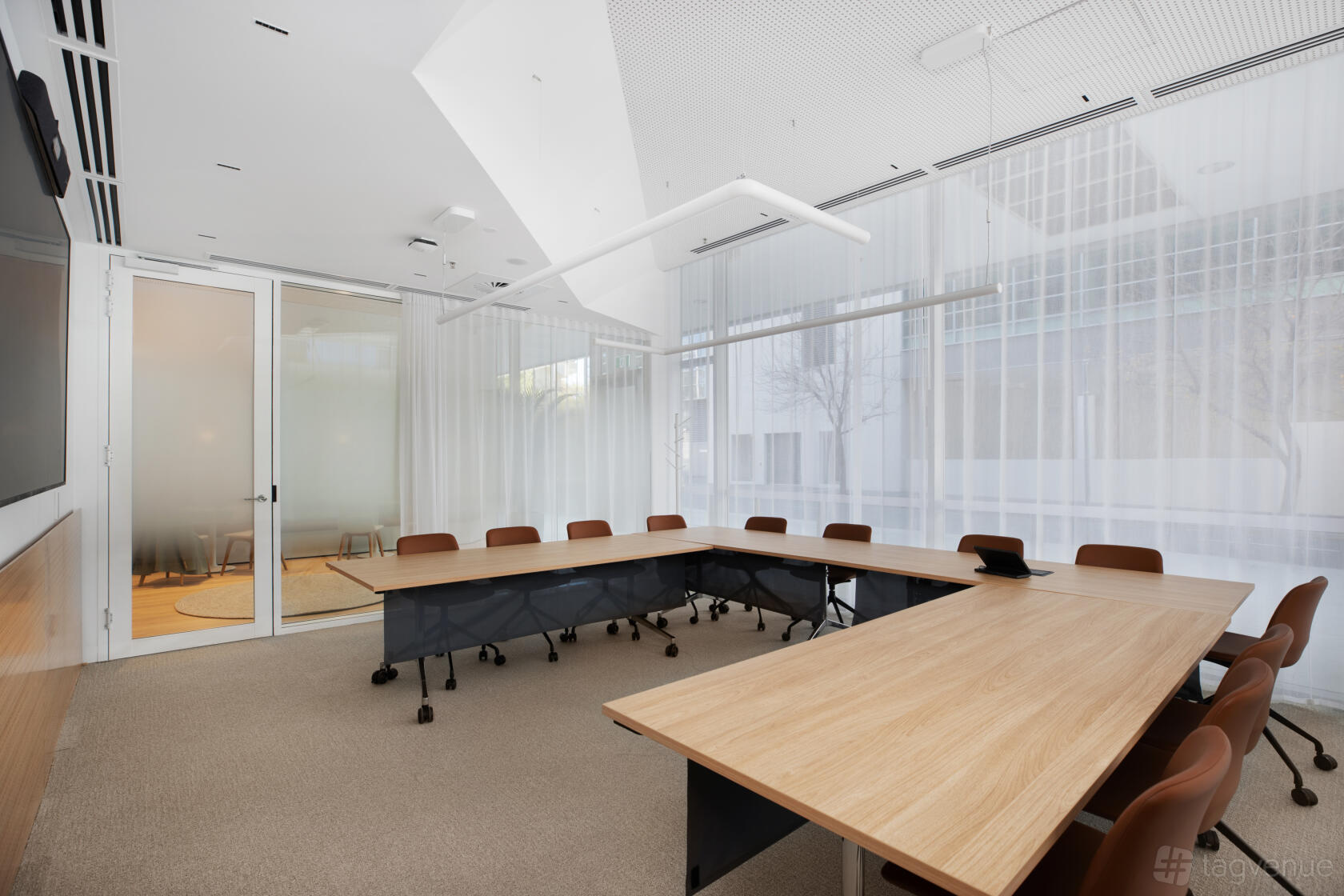 A training room with U-shaped wooden tables, brown chairs, floor-to-ceiling windows, and sheer curtains at Flex by ISPT Pathway Place.
