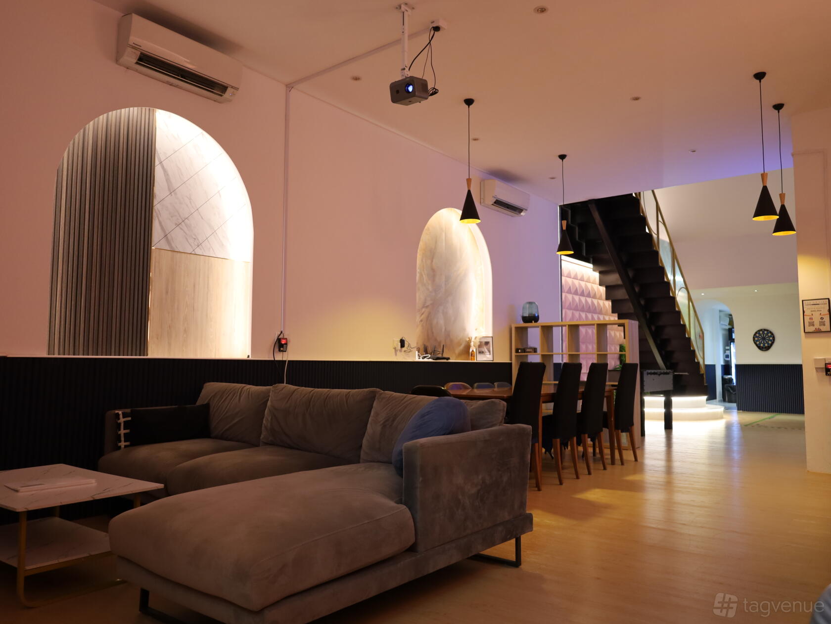 A function room with a grey sectional sofa, long dining table, pendant lighting, and staircase at All In One (Bendemeer).