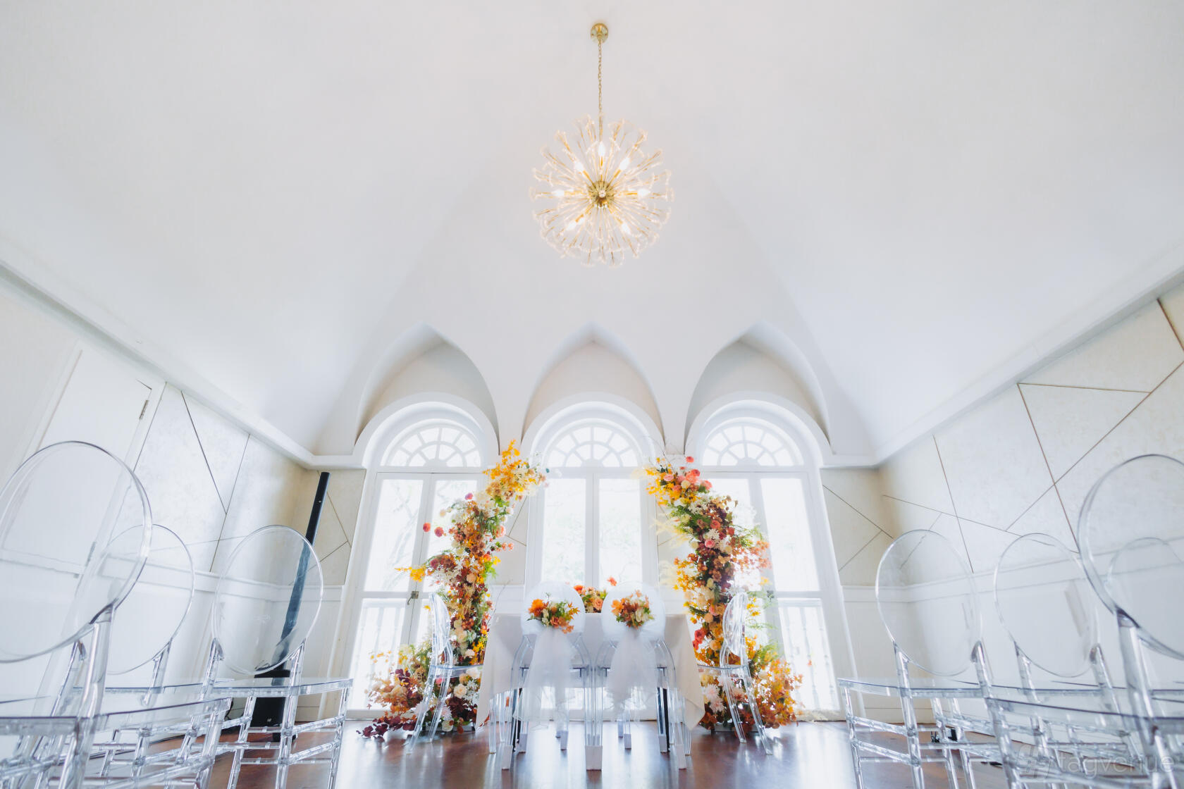 A function room with arched windows, floral arrangements, and transparent chairs at The Secret Patio.
