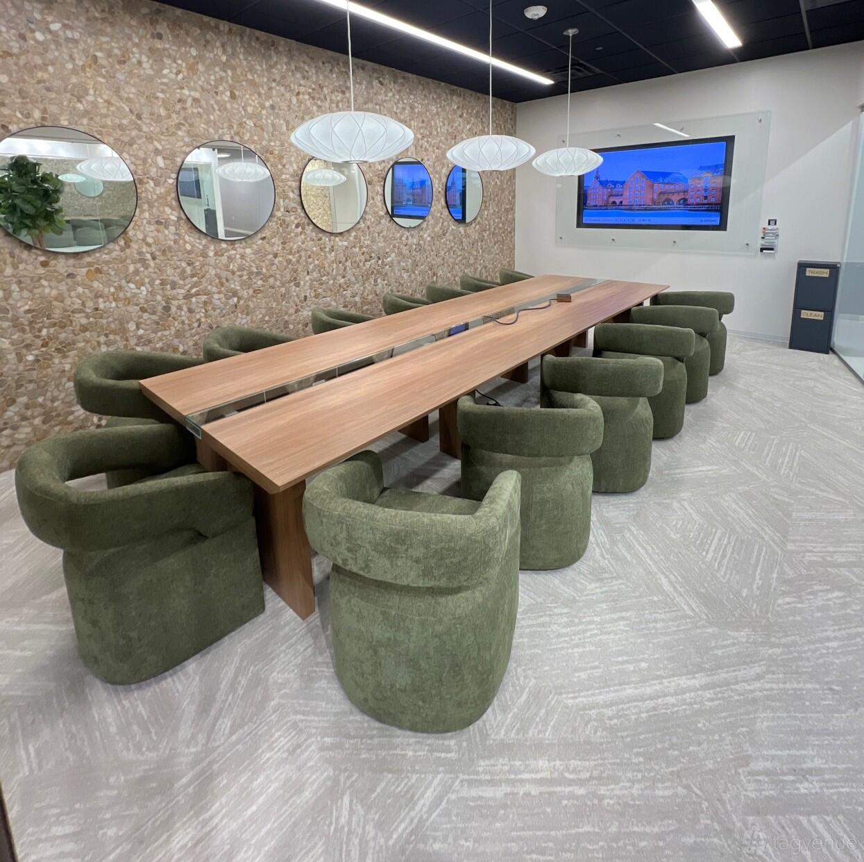 A boardroom with a long wooden table, green upholstered chairs, wall mirrors, and a mounted screen at Lucid Private Offices - McKinney/Craig Ranch.