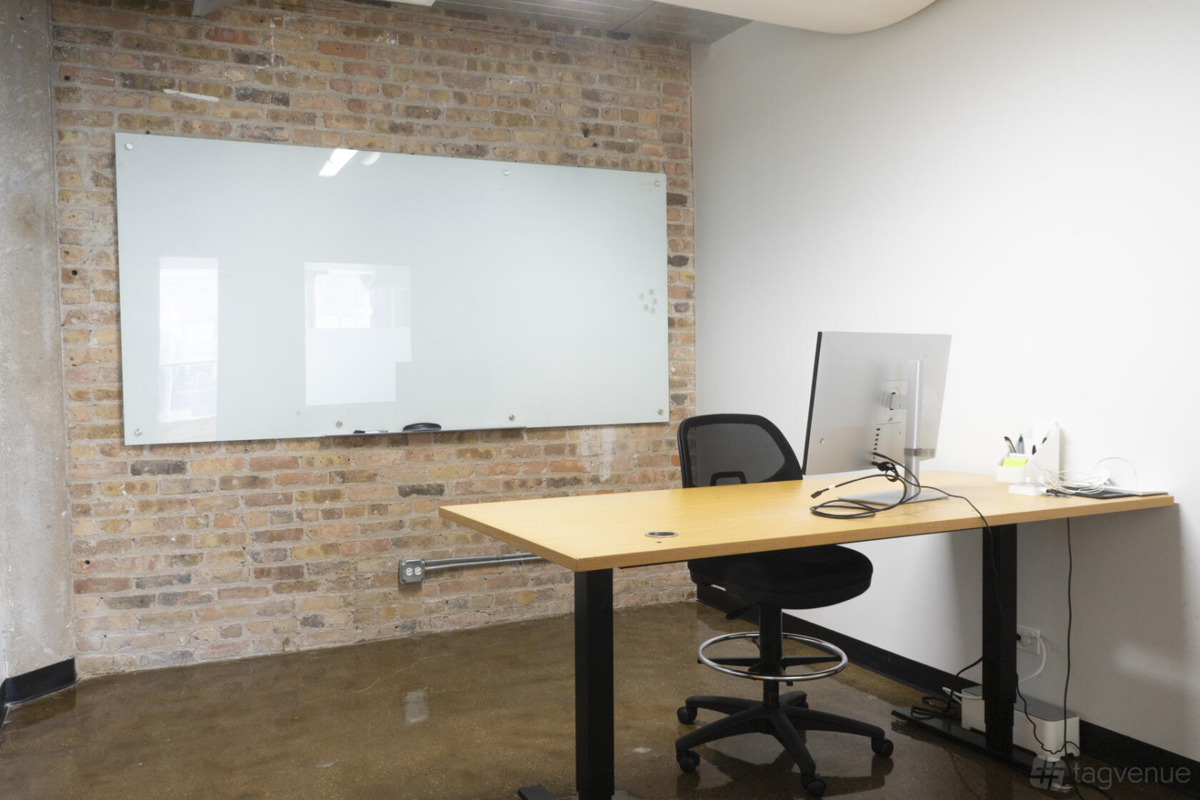 A meeting room with an exposed brick wall, glass whiteboard, and a desk with a computer at One Three Workspace.