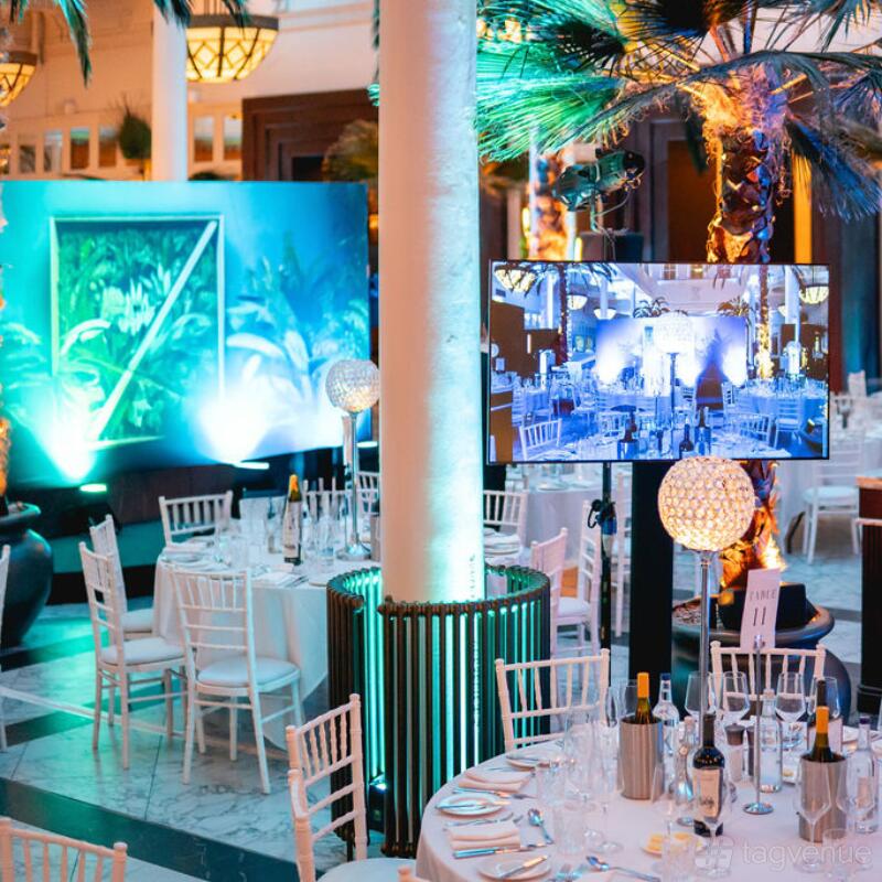 A hotel bar event space with white chairs, round tables, palm trees, and glowing blue accent lighting at Municipal Hotel Liverpool - MGallery.