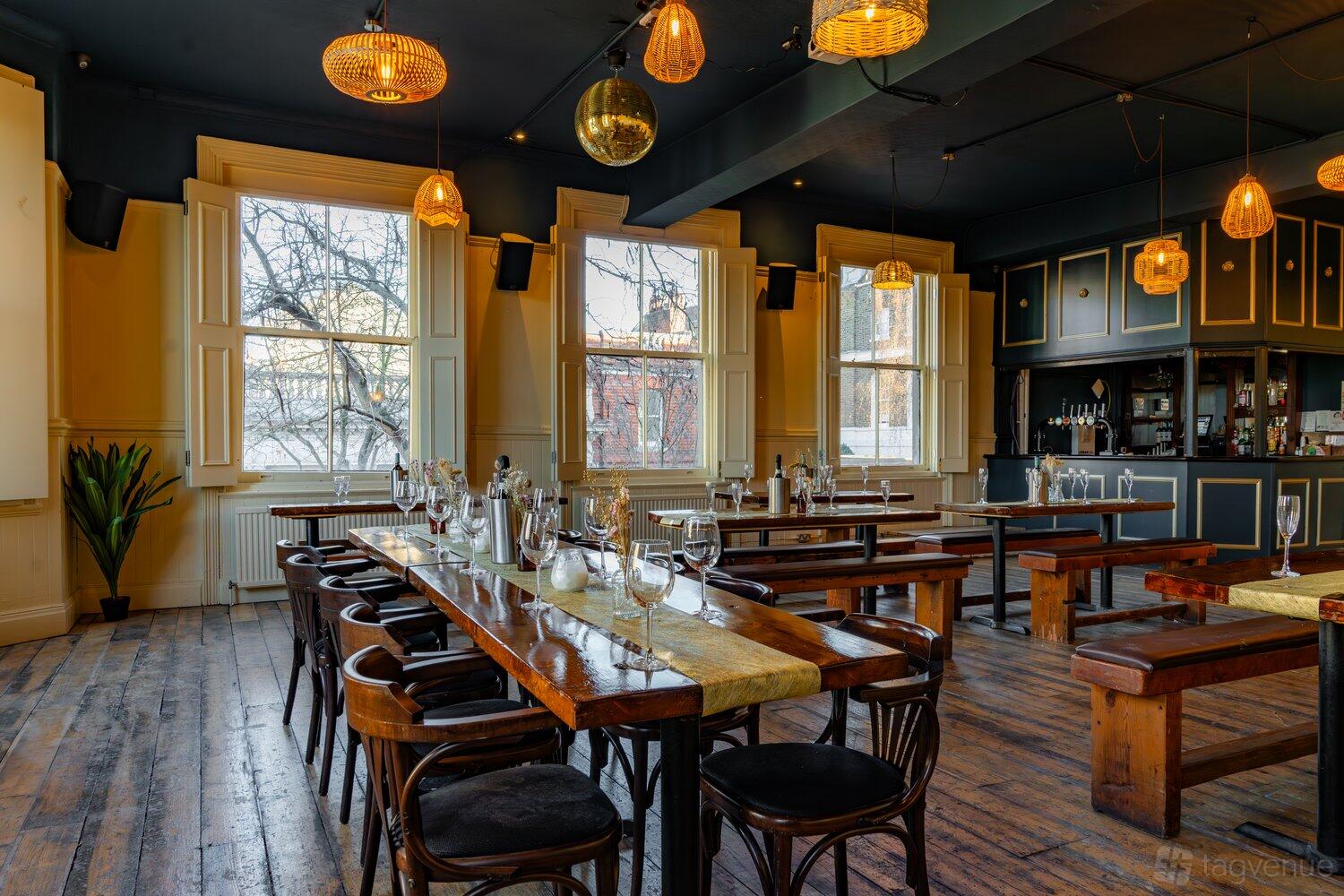 A function room in a pub with long wooden tables, black chairs, pendant lighting, and large windows at The Birds.