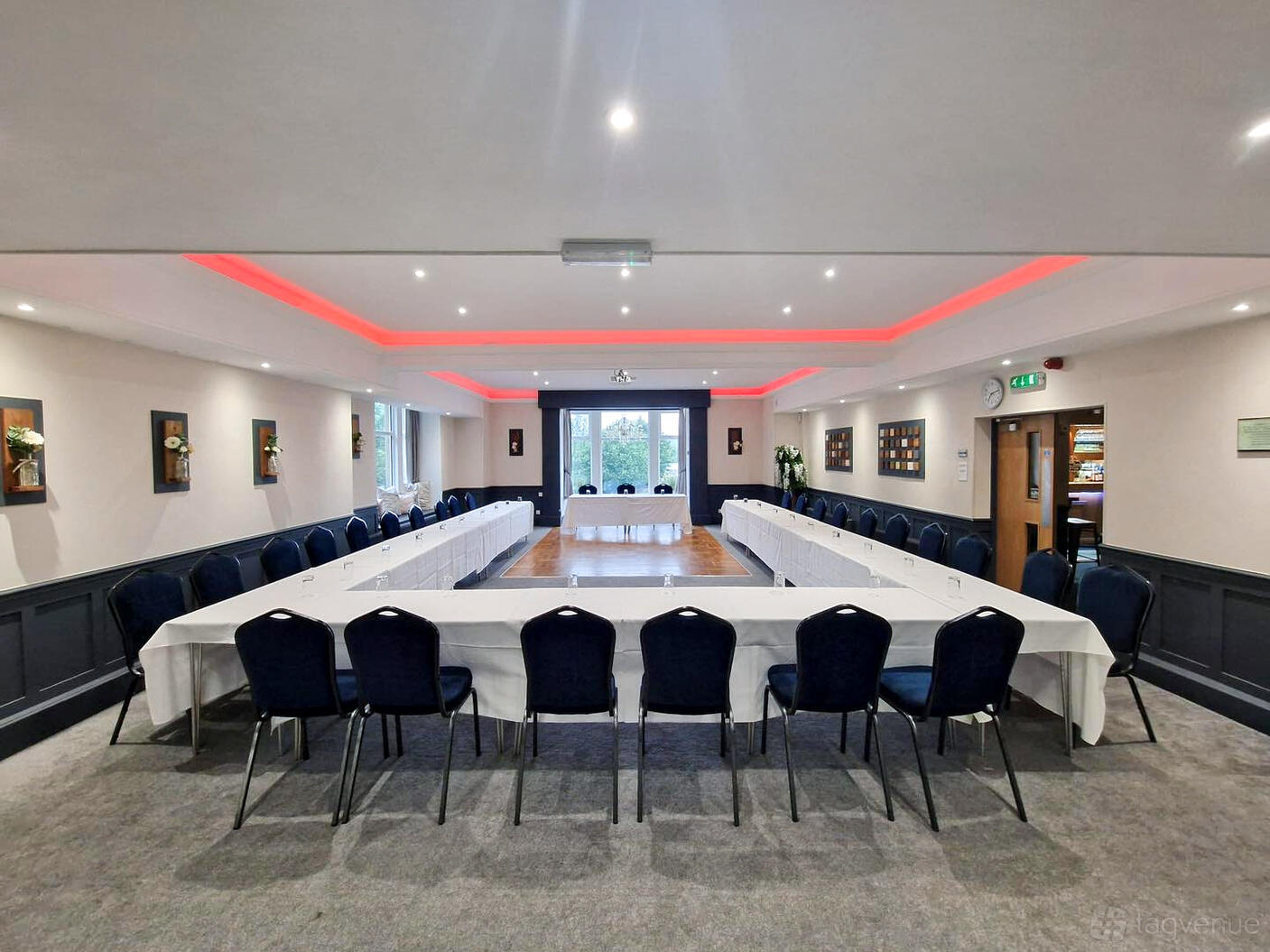 A function room with U-shaped tables covered in white linens, navy chairs, and accent lighting at The Mill House.