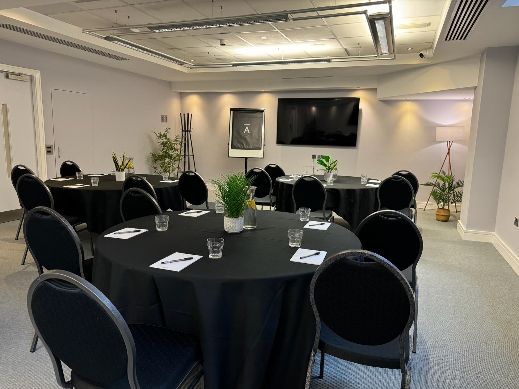 A meeting room with round tables, notepads, black chairs, a large screen, and potted plants at The Alan.