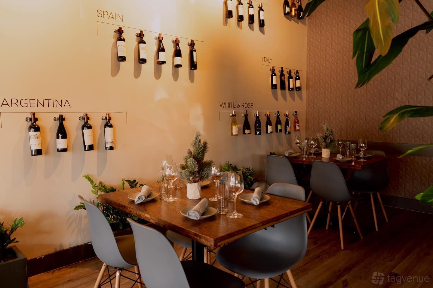 A dining room with wall-mounted wine displays labeled by region and wooden tables set for guests at Reunion Ktchn Bar.