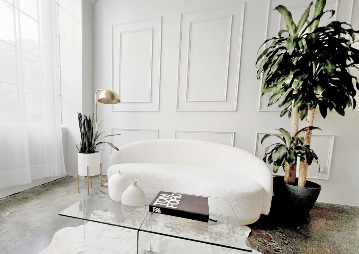 A photo studio with white paneled walls, a curved white sofa, tall plants, and sheer curtains at Vera Lofts | Vera.
