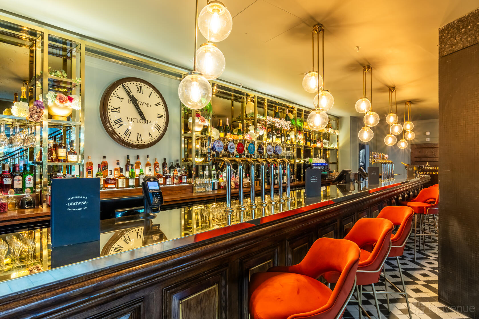 A bar in a restaurant with mirrored shelves, globe pendant lights, and orange velvet stools at Browns Victoria.