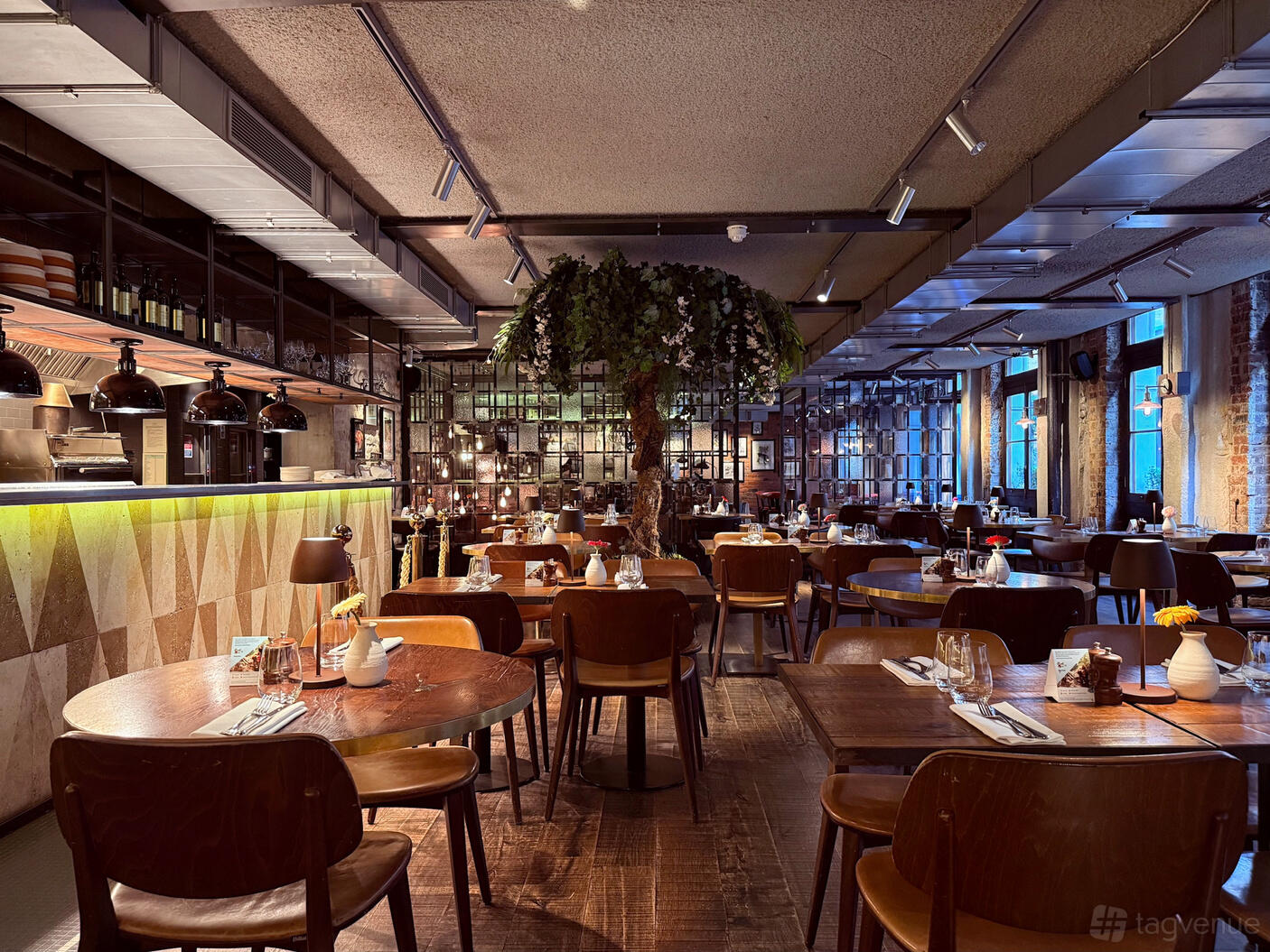 A restaurant with wooden tables, brown chairs, a central tree feature, and floor-to-ceiling windows at Canto Corvino.