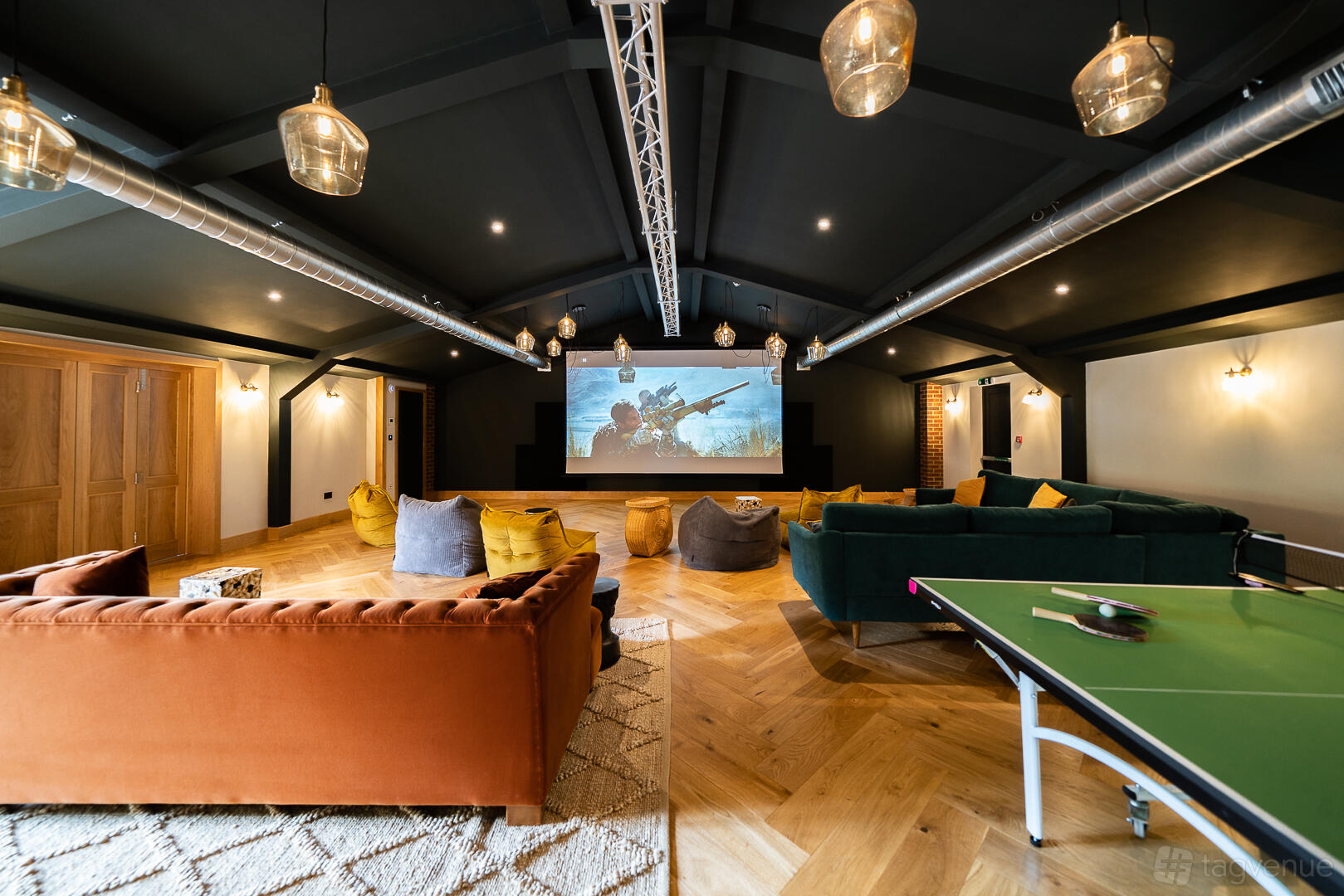 A hotel screening room with comfortable sofas, bean bags, wood flooring, and a large projector screen at Tewinbury Farm Hotel.