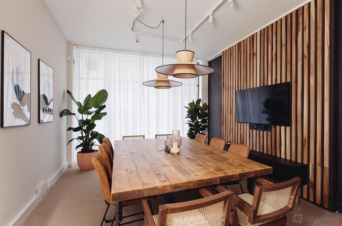 A meeting room with a wooden conference table, rattan chairs, pendant lights, and a wall-mounted screen at Vine Hill, Farringdon.