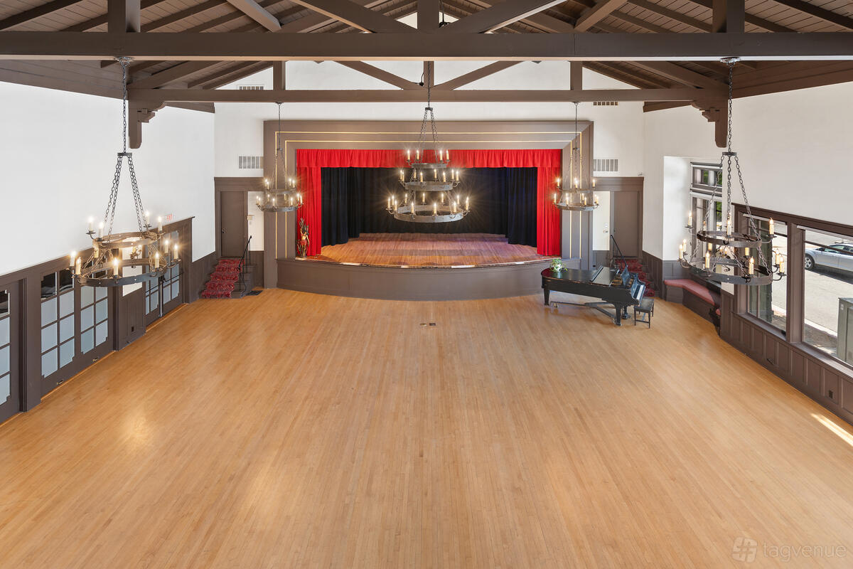 A hall in a historic building with exposed beams, polished wood floors, chandeliers, and a stage at The South Pasadena Clubhouse.