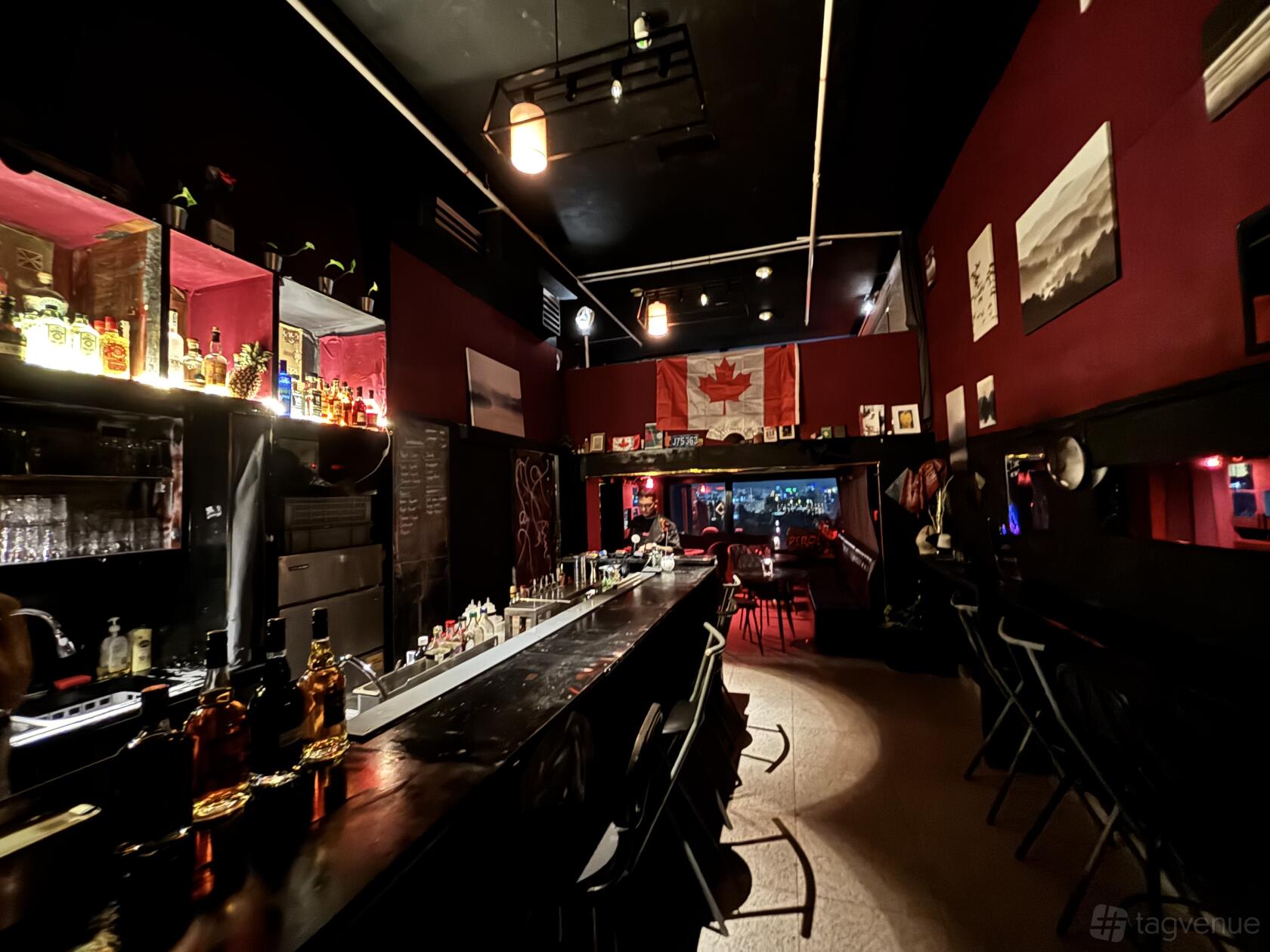 A cocktail bar with a long black counter, red walls, shelves of liquor, and a Canadian flag at The Cocktail Office.