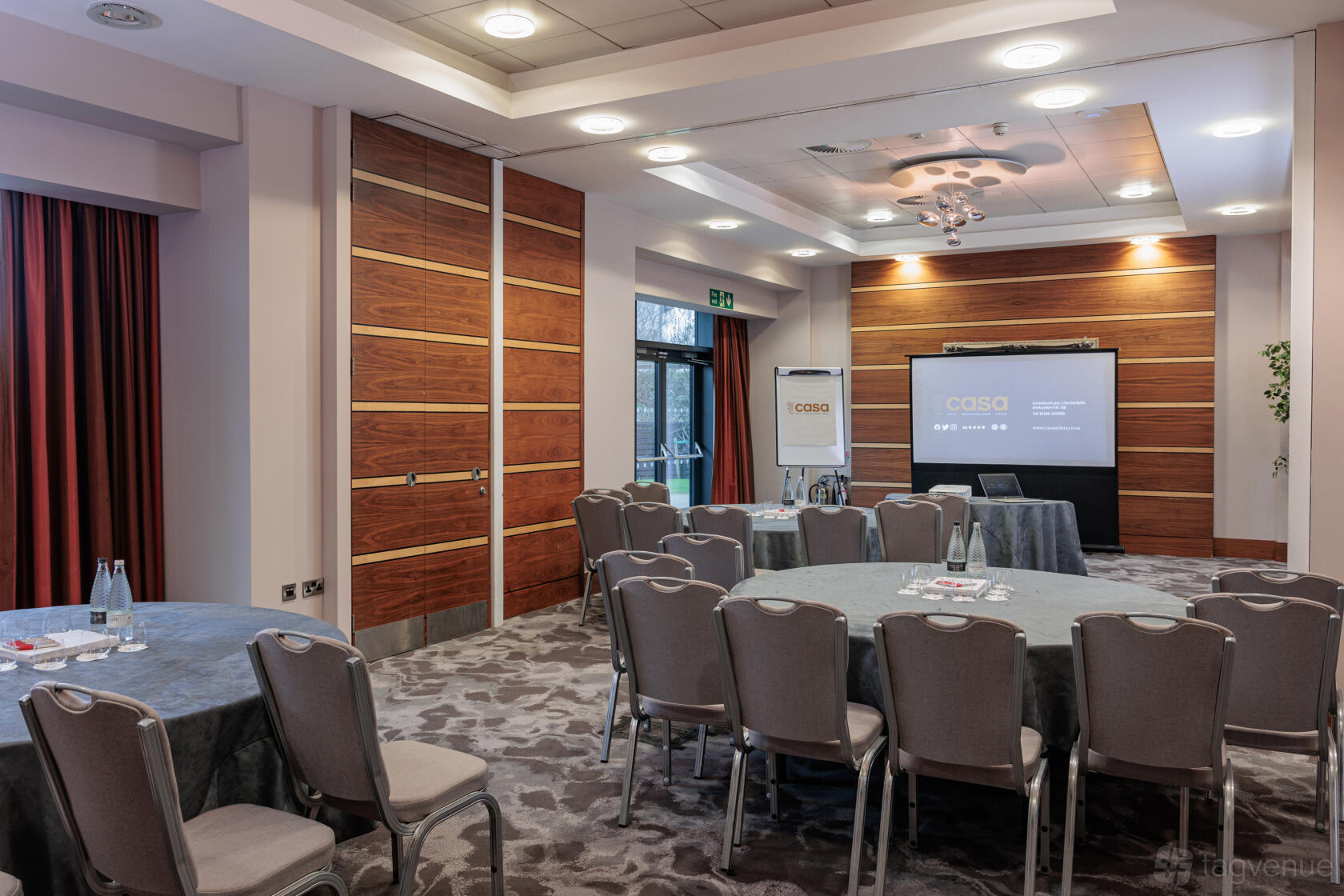 An event space with round tables, padded chairs, wood paneling, and a projector screen at Casa Hotel.