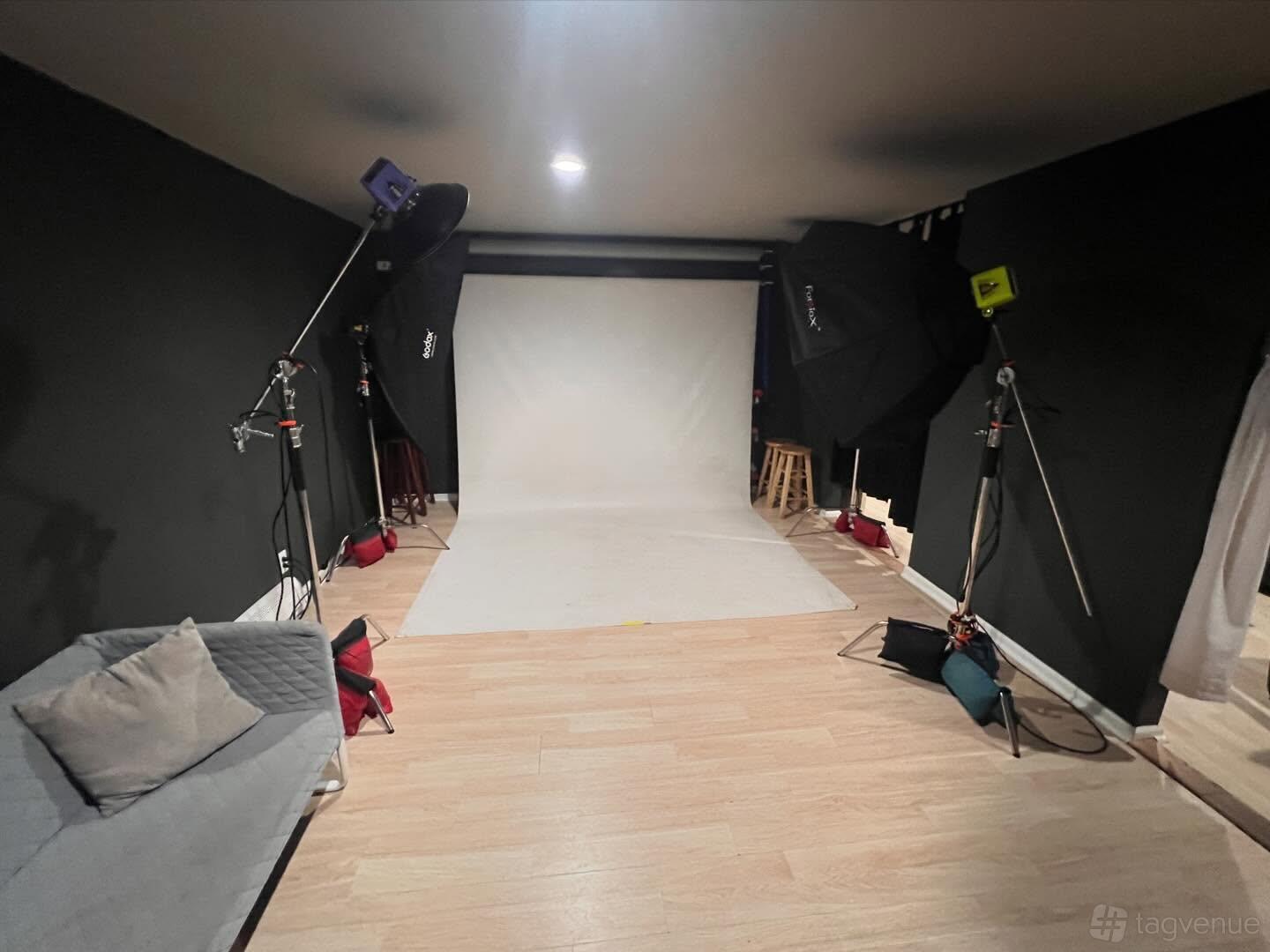 A photo studio with a white backdrop, softbox lights, wooden floors, and a grey sofa at Studio 210.