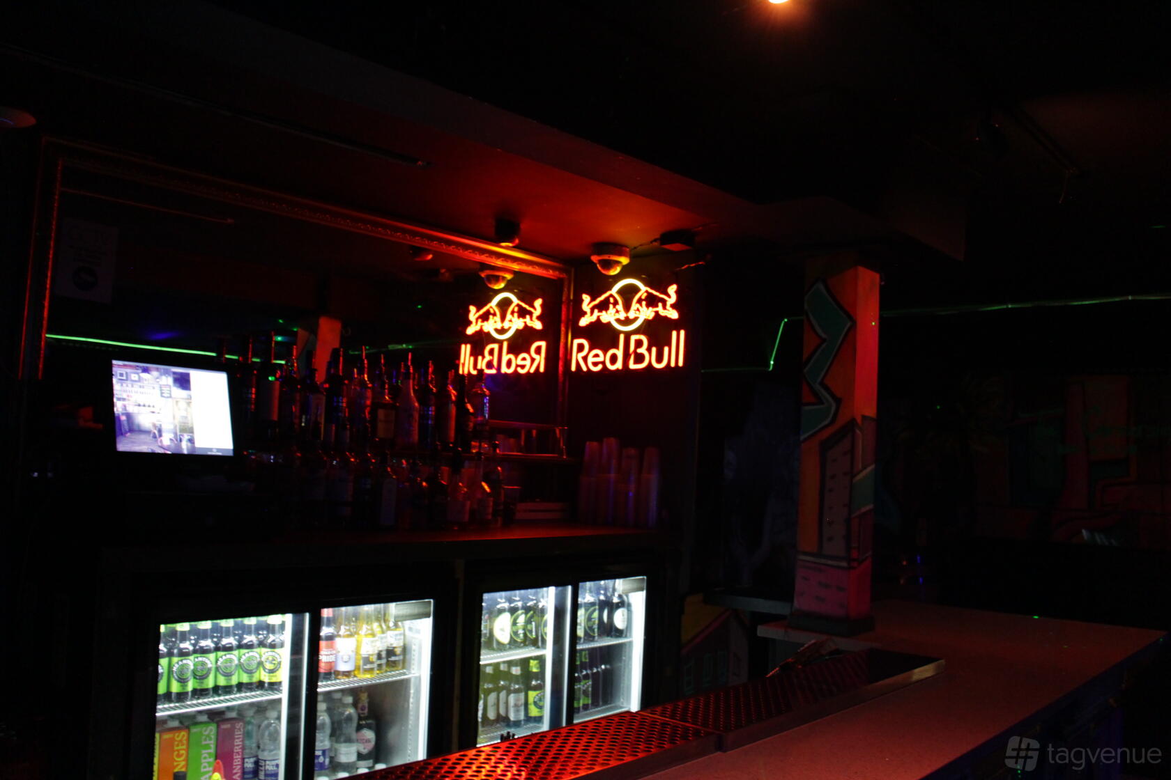 A basement bar with neon Red Bull signage, stocked bottles, and under-counter fridges at The Cornershop Bar.