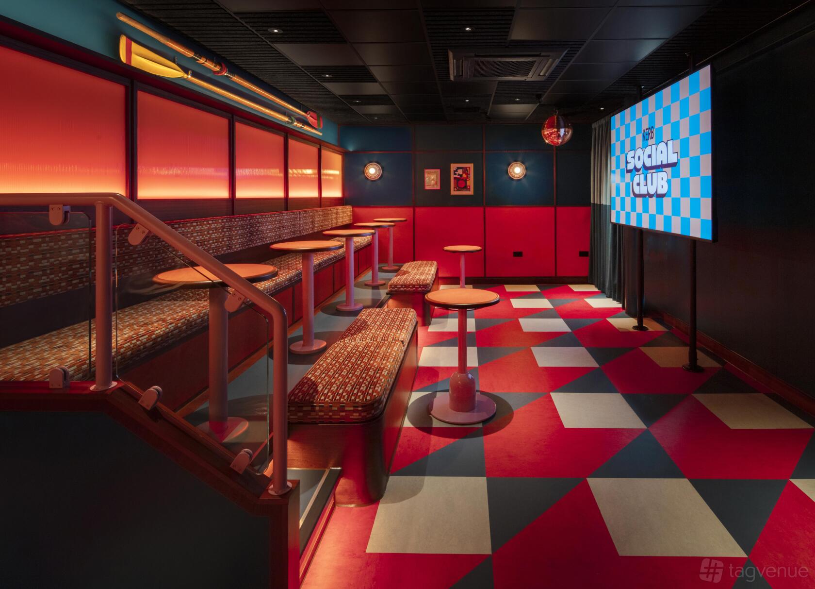 An event space in a bar with checkered flooring, stadium-style seating, and a large projector screen at KERB Social Club Spitalfields.