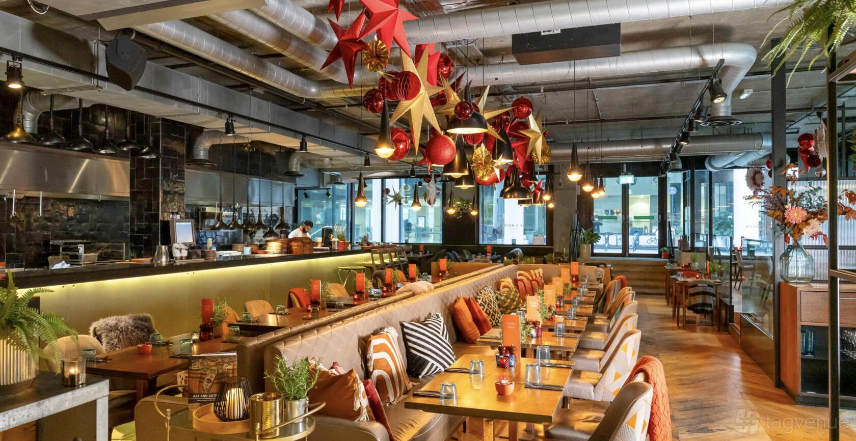 A bar with eclectic pendant lights, exposed ductwork, festive hanging ornaments, and banquette seating at Drake & Morgan at King's Cross.