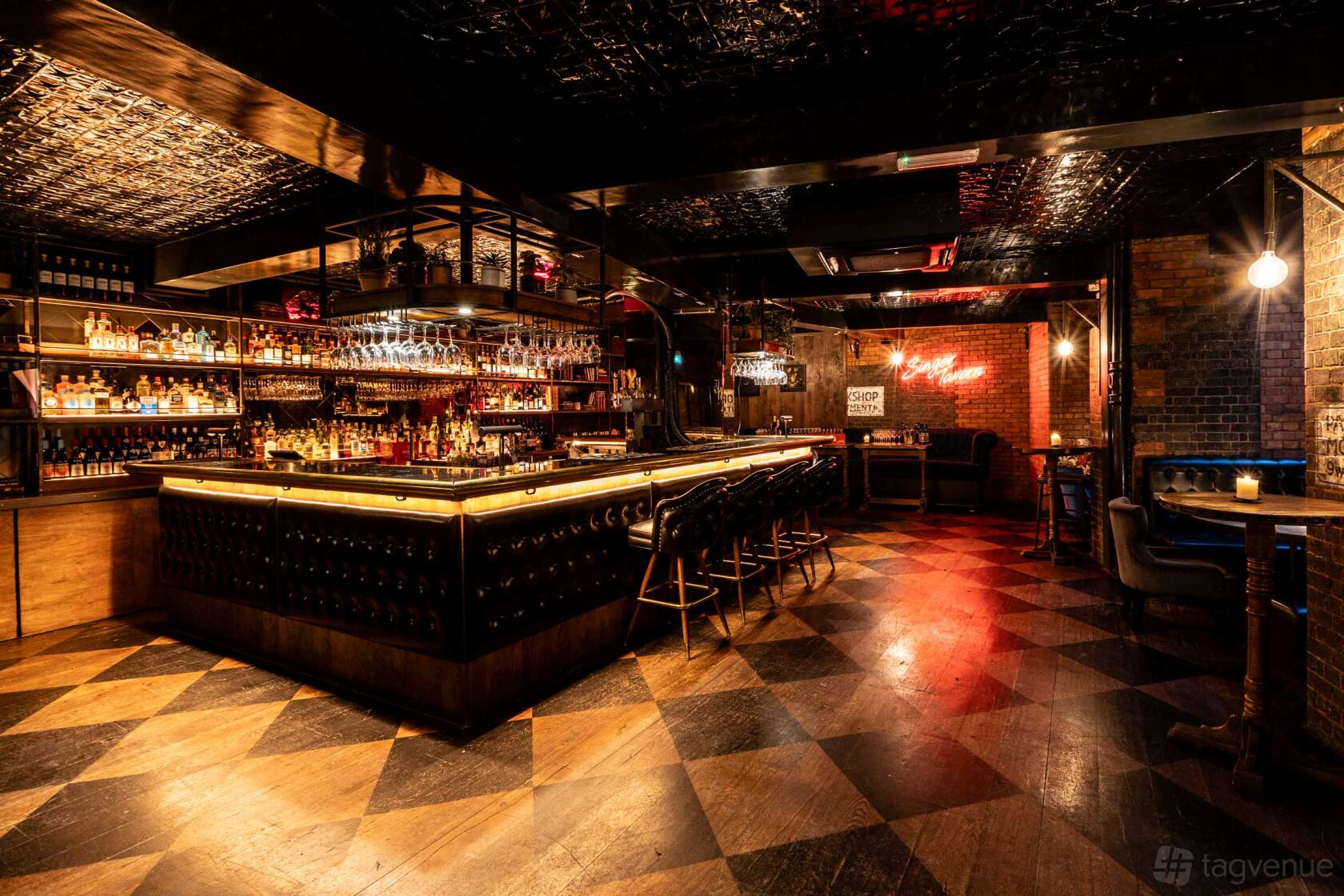 A bar with checkered wood flooring, a central counter lined with stools, and shelves of bottles at Singer Tavern 5CC Bar.