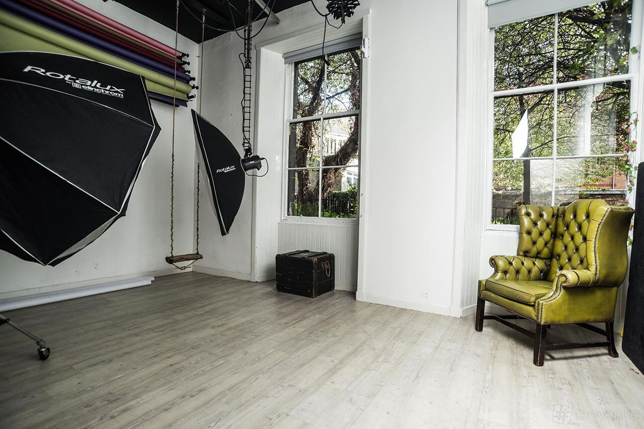 A photo studio with seamless backdrop rolls, softbox lighting, a vintage green chair, and large windows at Nicola Bald Photography.