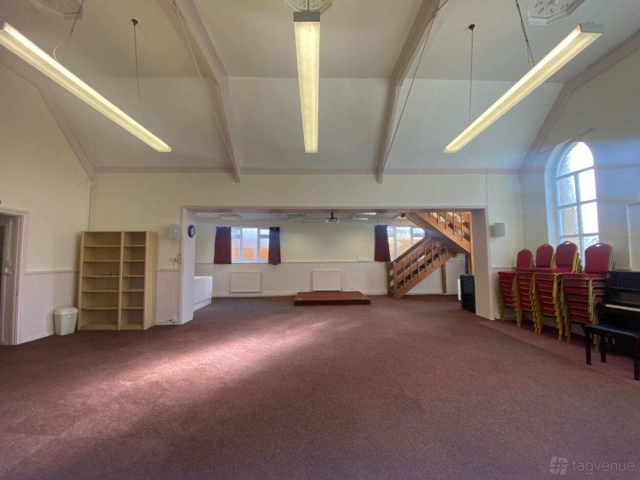 A spacious event hall with carpeted floors, high ceilings, a small raised stage, and large windows at Foulsham Hall.