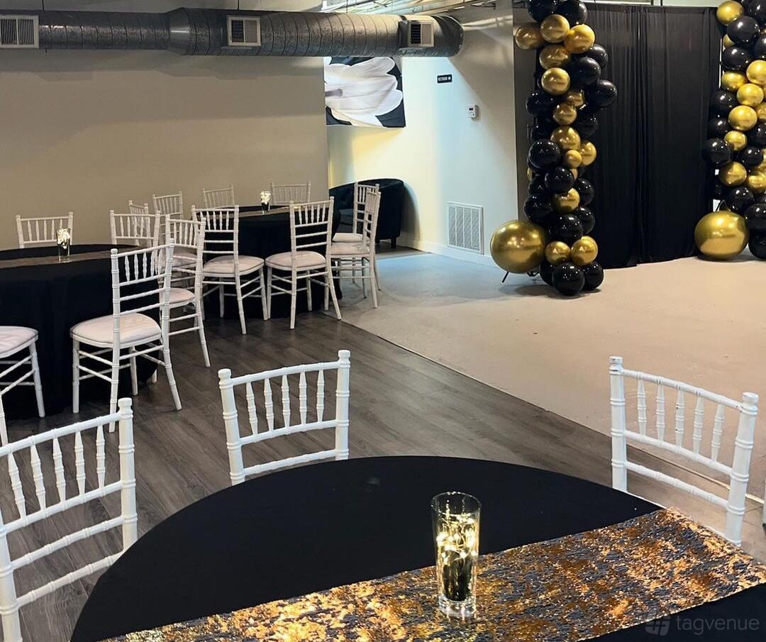 An event venue with white chairs, round tables with black linens, and gold and black balloon columns at Oznorras Event Space.