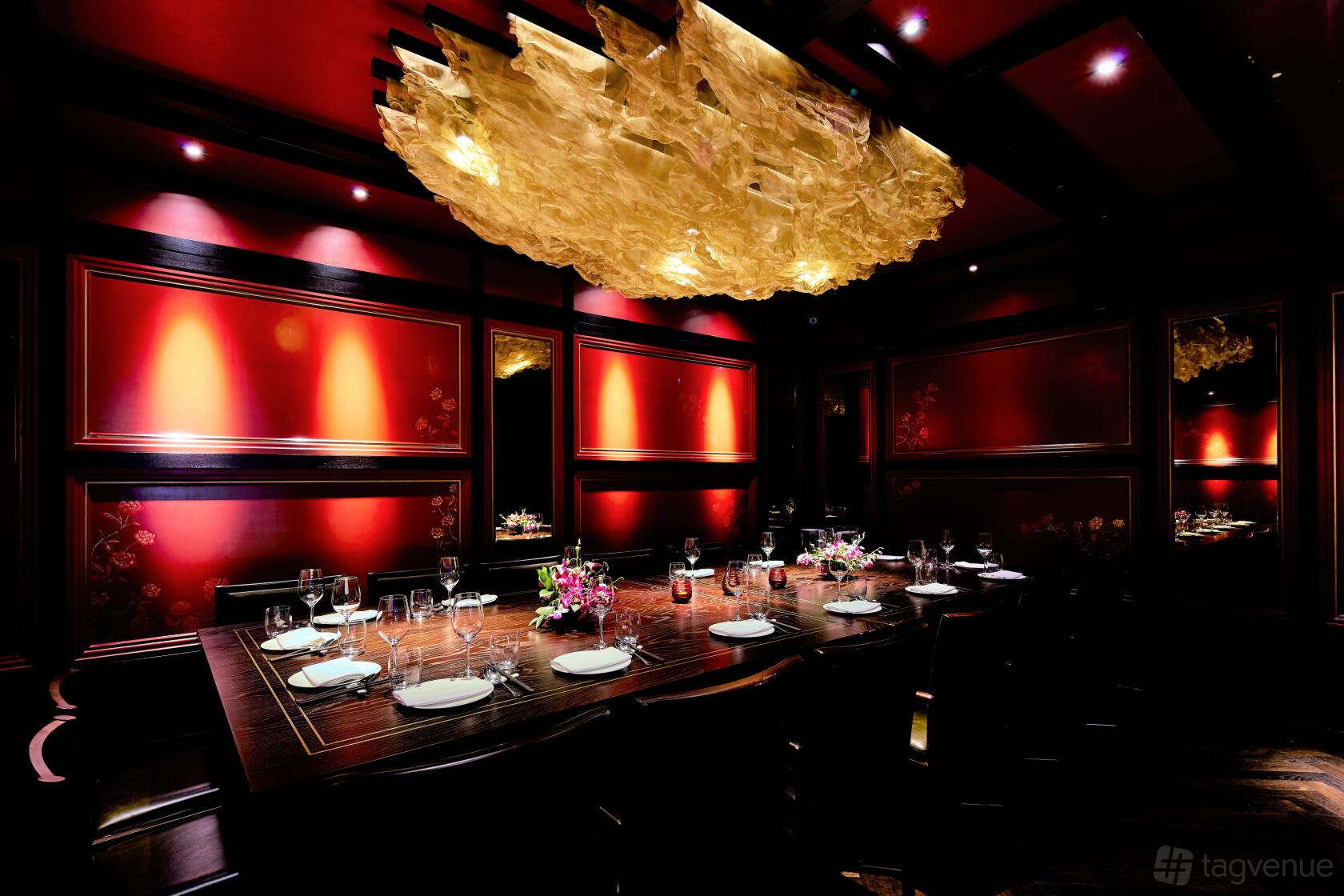 A restaurant dining room with dark wood paneling, red lighting, and a sculptural gold ceiling fixture at Hakkasan Mayfair.