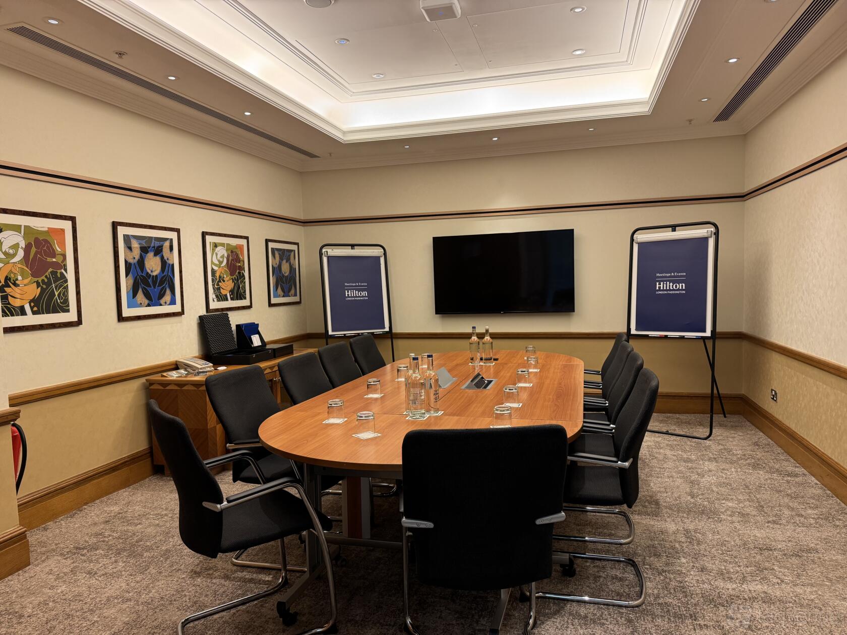 A boardroom with a wooden oval table, black chairs, wall art, and a large screen at Hilton London Paddington.
