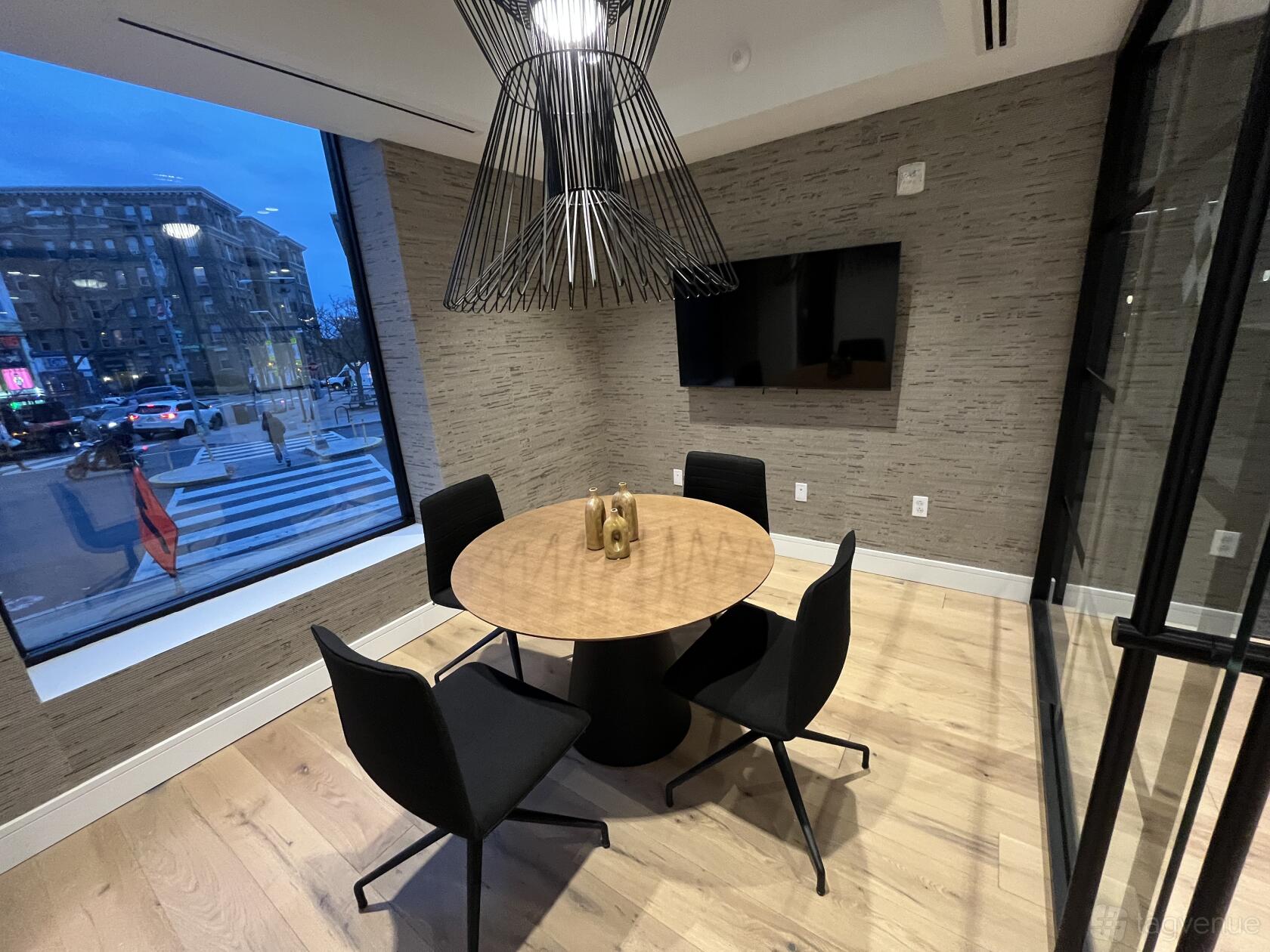 A meeting room with a round wooden table, four black chairs, large windows, and a wall-mounted TV at Venture X Washington DC - Adams Morgan.