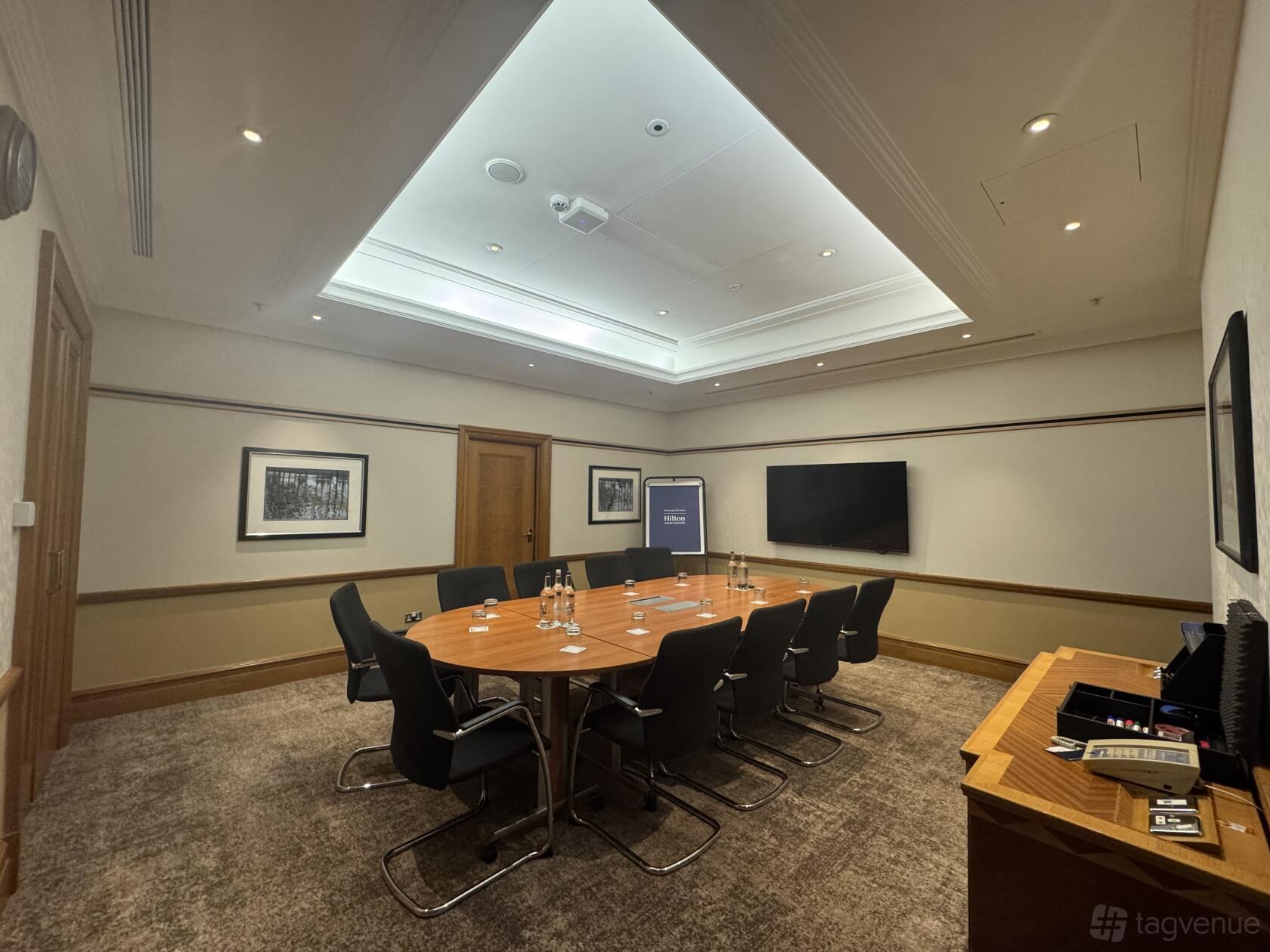 A boardroom with a rectangular wooden table, executive chairs, wall-mounted screen, and framed art at Hilton London Paddington.