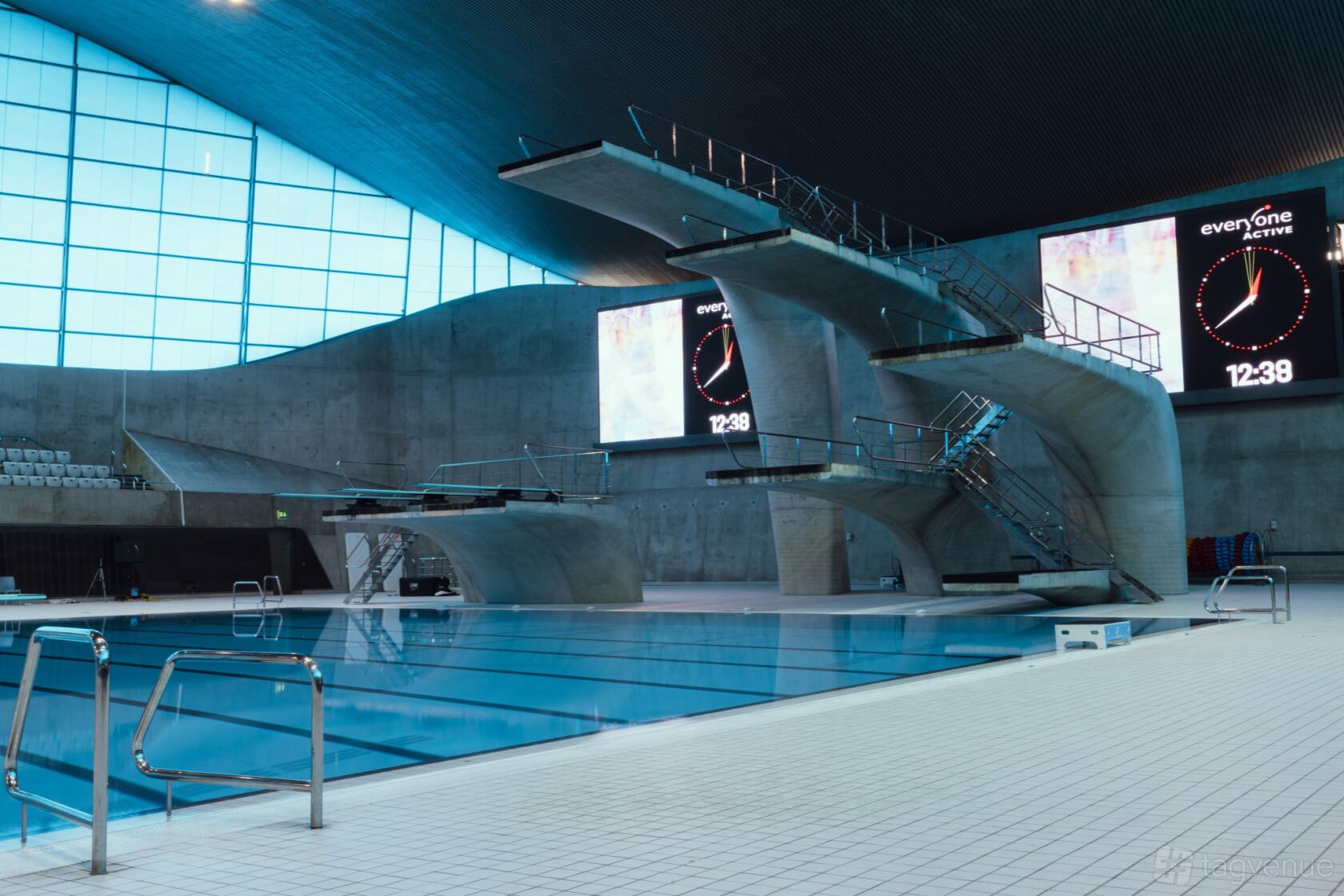 A swimming pool with concrete diving platforms and digital clocks at London Aquatics Centre.