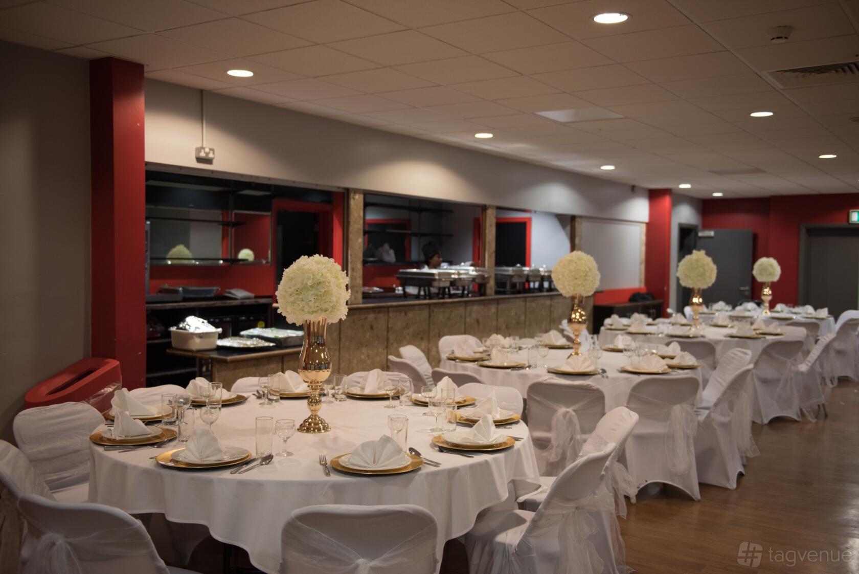 A banquet hall with round tables draped in white linens, gold centerpieces, and buffet counters at Harrow Leisure Centre.