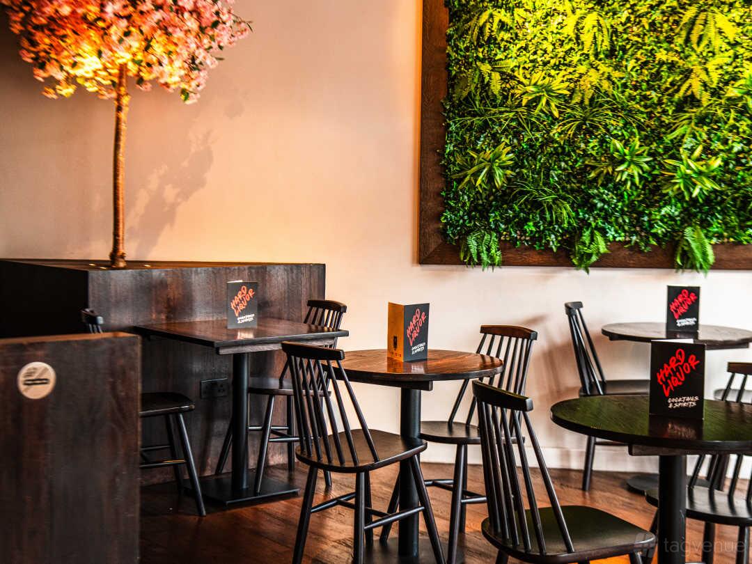 A restaurant with round wooden tables, black chairs, a wall of greenery, and a flowering decorative tree at Trap Kitchen.
