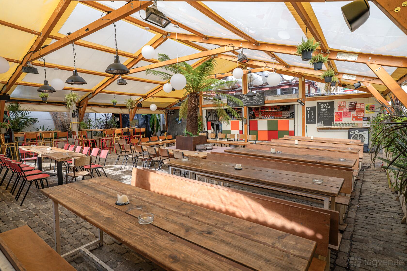 A bar with long wooden tables, mixed seating, hanging lights, and a glass canopy at Peckham Arches.