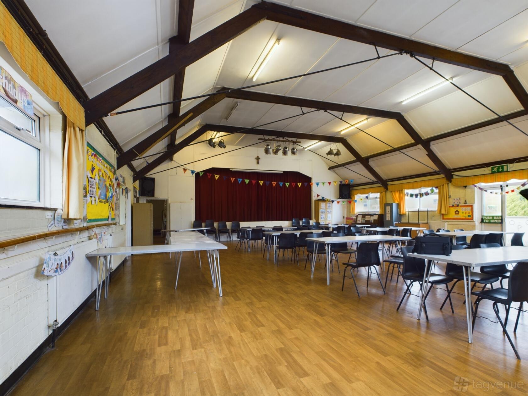 An event space with wooden floors, exposed beams, foldable tables, and a stage with red curtains at St. Clement's Church Hall.