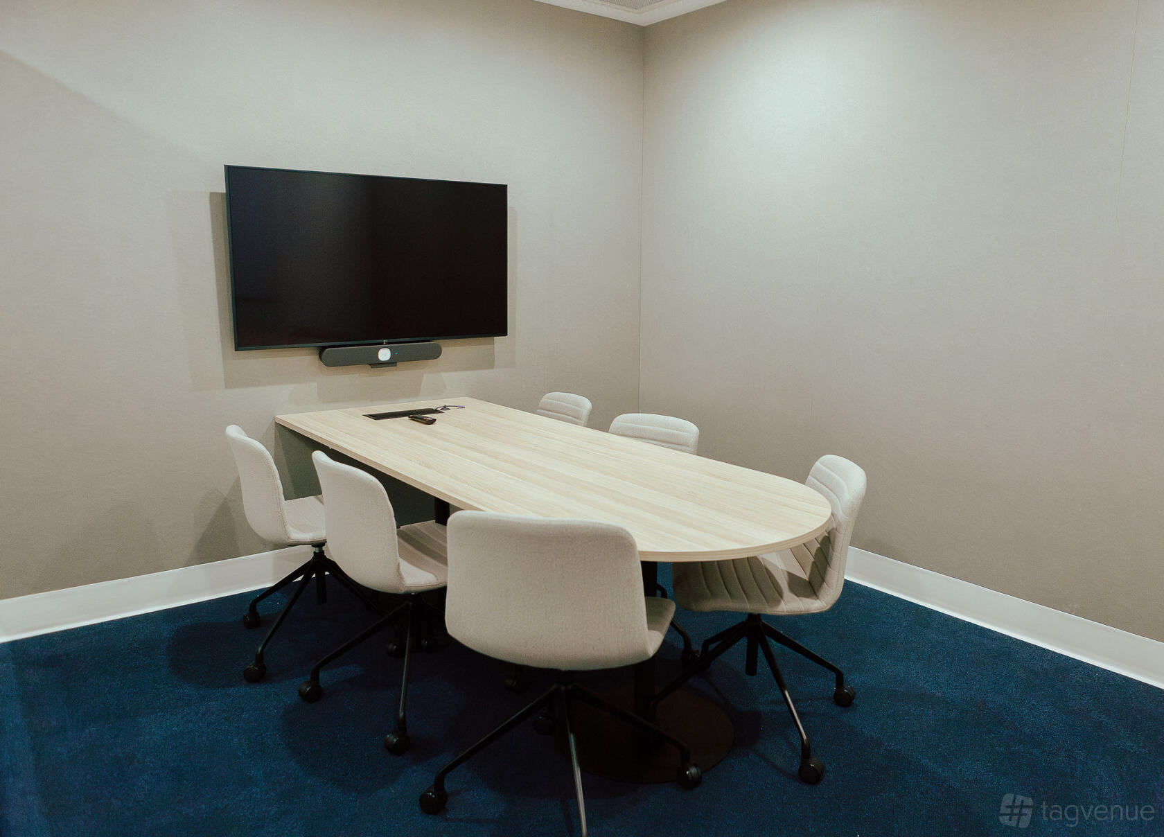 A meeting room with a light wood oval table, six white office chairs, and a wall-mounted screen at Tank Stream Suites.