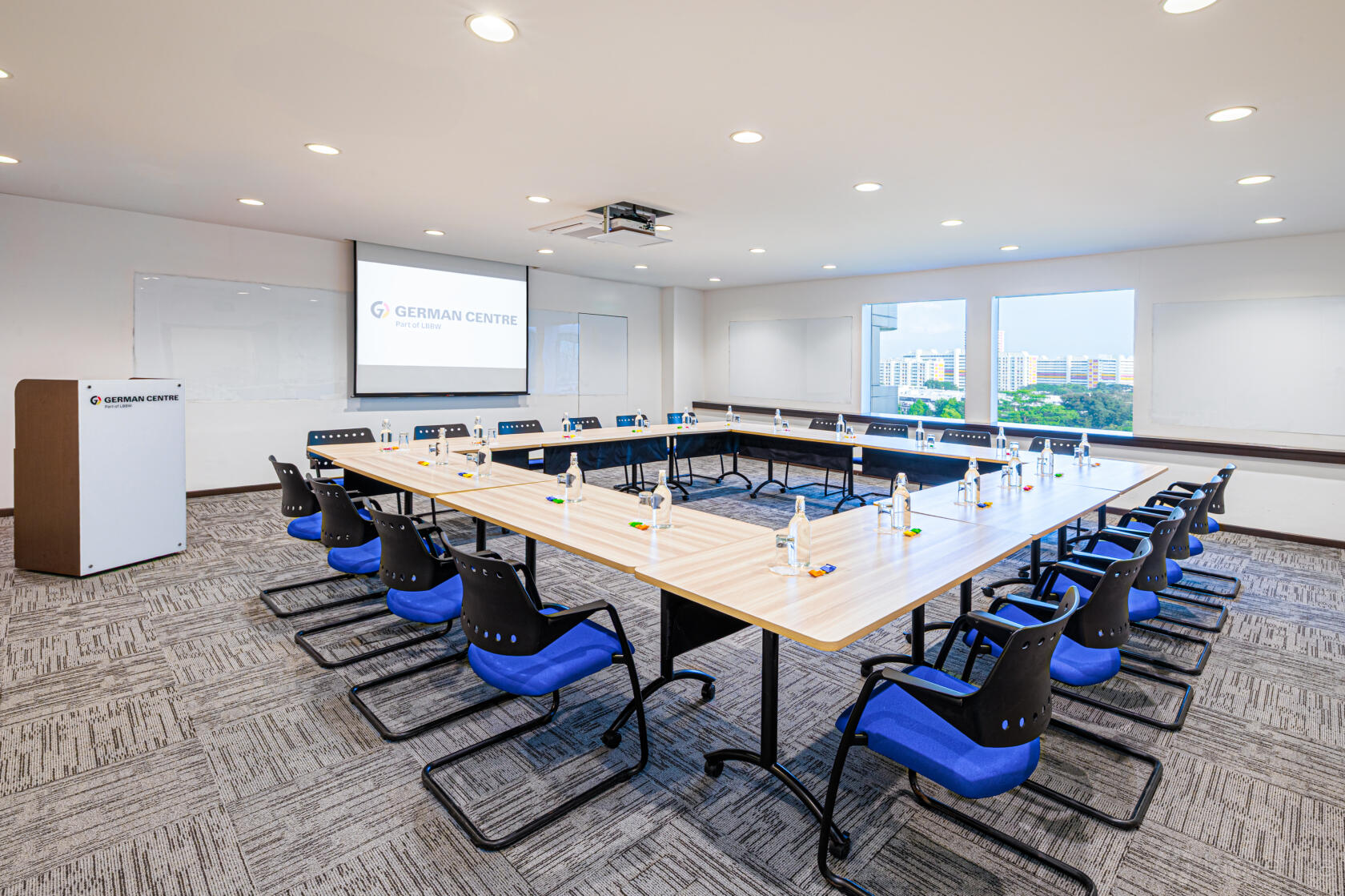 An auditorium with U-shaped tables, blue chairs, bottled water, and large windows at German Centre Singapore.