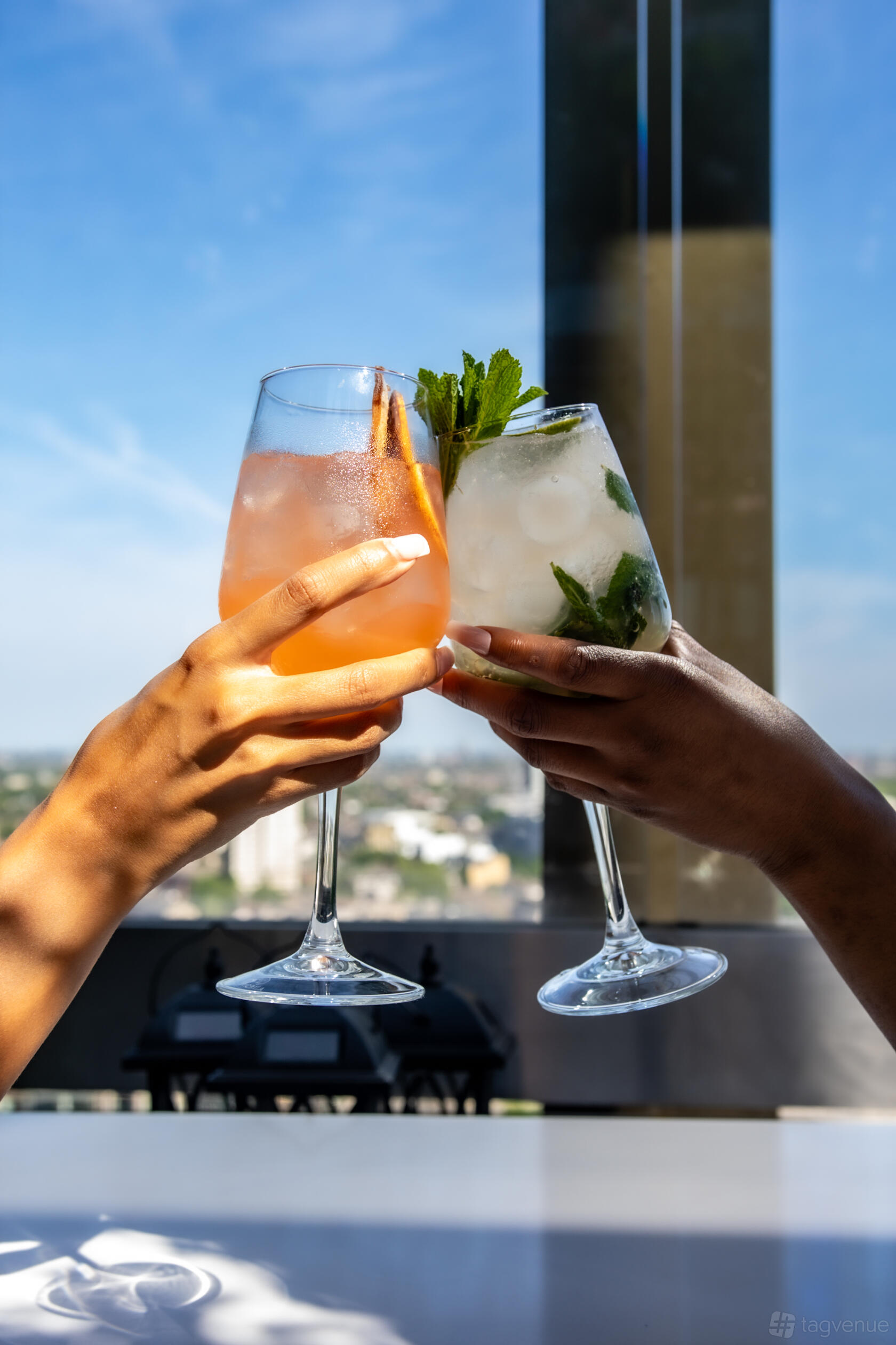 A restaurant setting with two hands holding cocktails against a blurred city skyline at STK Rooftop - Stratford.