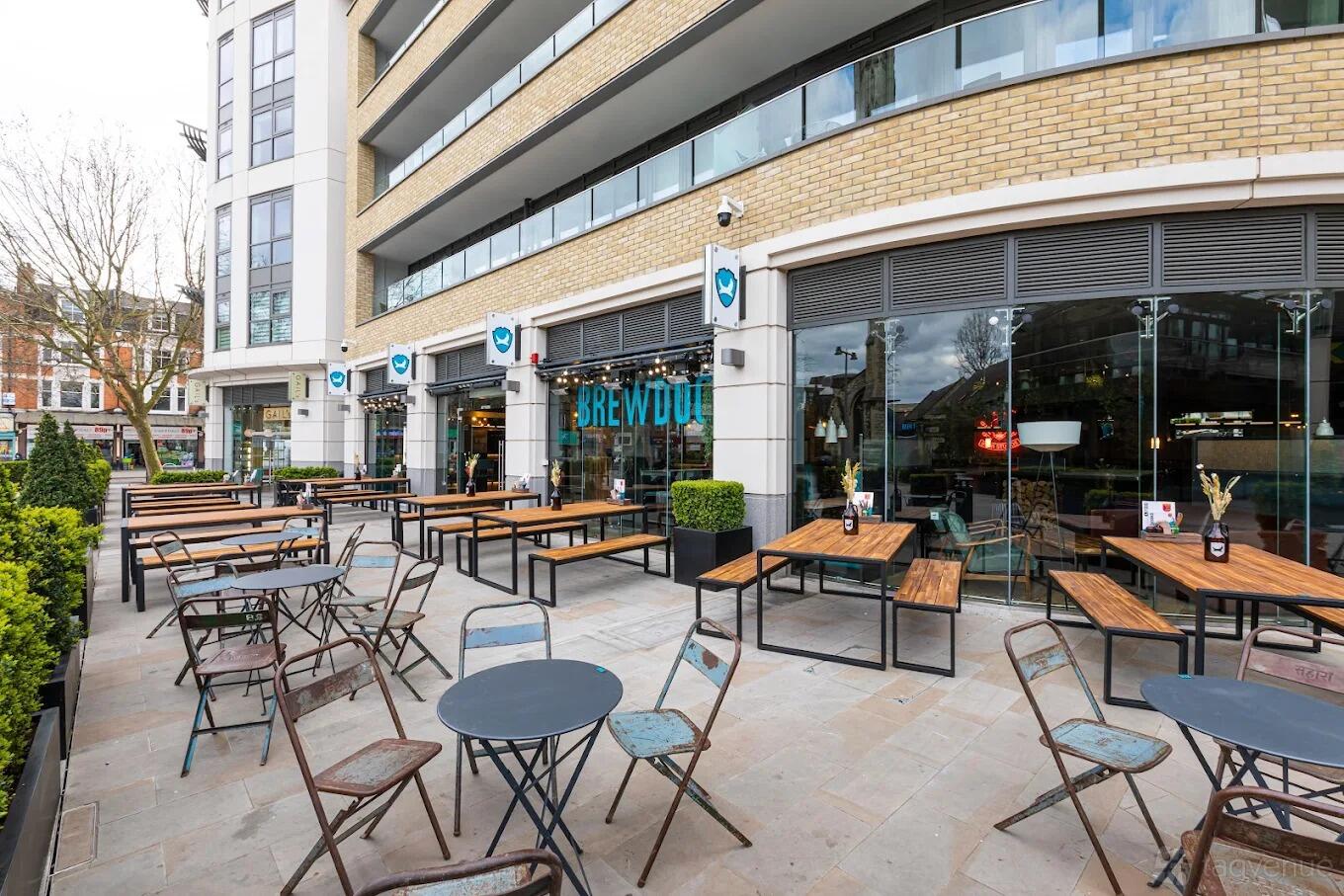 An outdoor bar area with wooden picnic tables, metal chairs, and potted plants at BrewDog Ealing.
