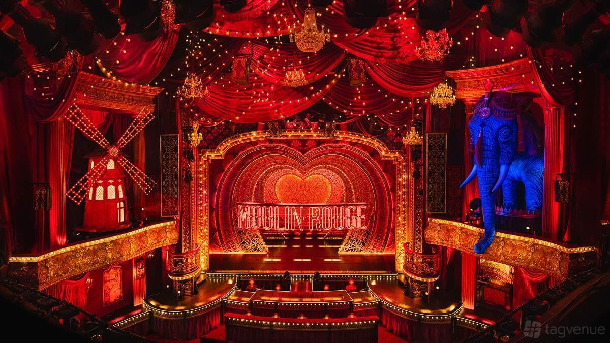 A theatre with red velvet drapes, ornate chandeliers, a lit windmill, and a heart-shaped Moulin Rouge stage at ATG VIP: Moulin Rouge - Piccadilly Theatre.