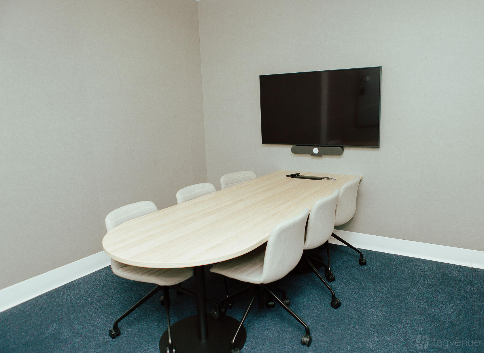 A coworking meeting room with a light wood table, six chairs, and wall-mounted screen at Tank Stream Suites.